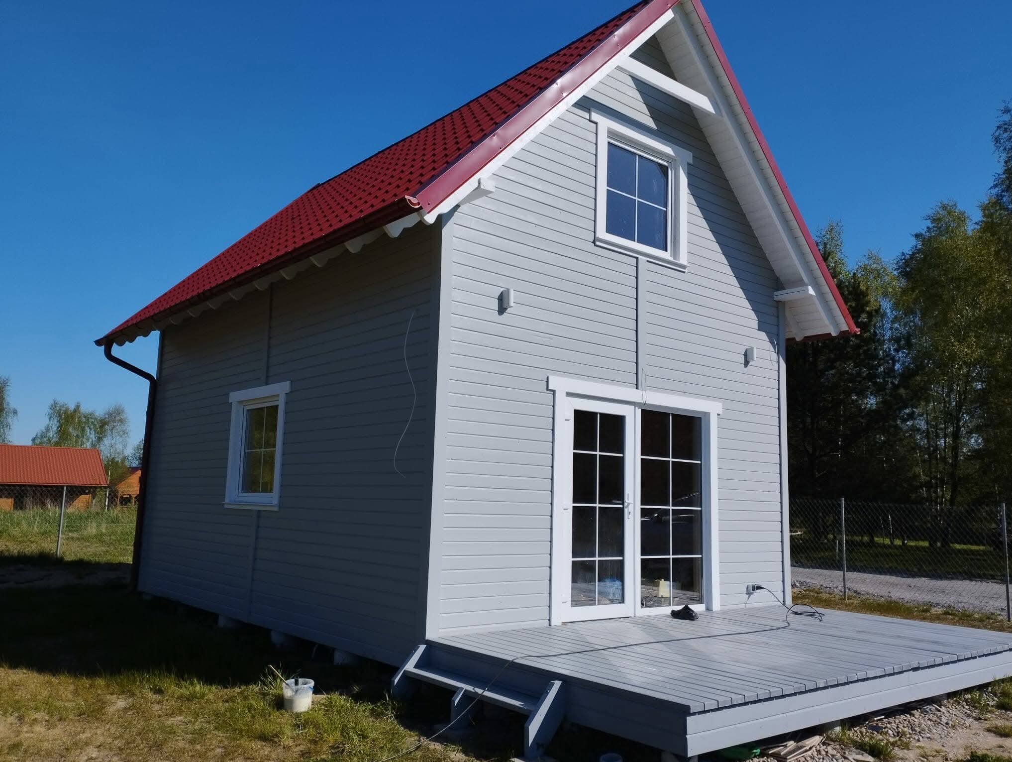 Szary dom szkieletowy z czerwonym dachem i drewnianym tarasem. Widoczne okna, drzwi balkonowe i wiadro z farbą. Błękitne niebo w tle.
