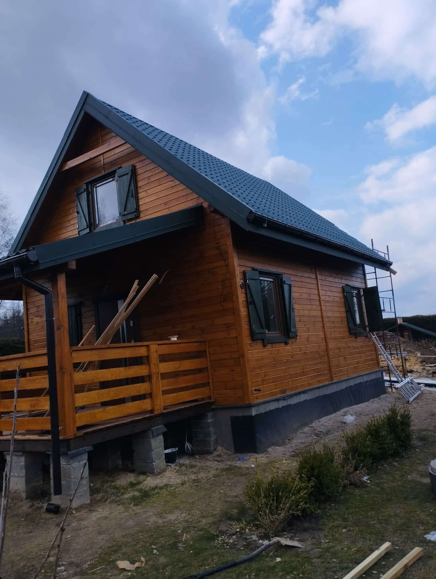 Drewniany dom szkieletowy z ciemną dachówką i okiennicami, widoczny z boku, z elementami rusztowania i przygotowanymi deskami, sugerujący etap budowy lub wykończenia.