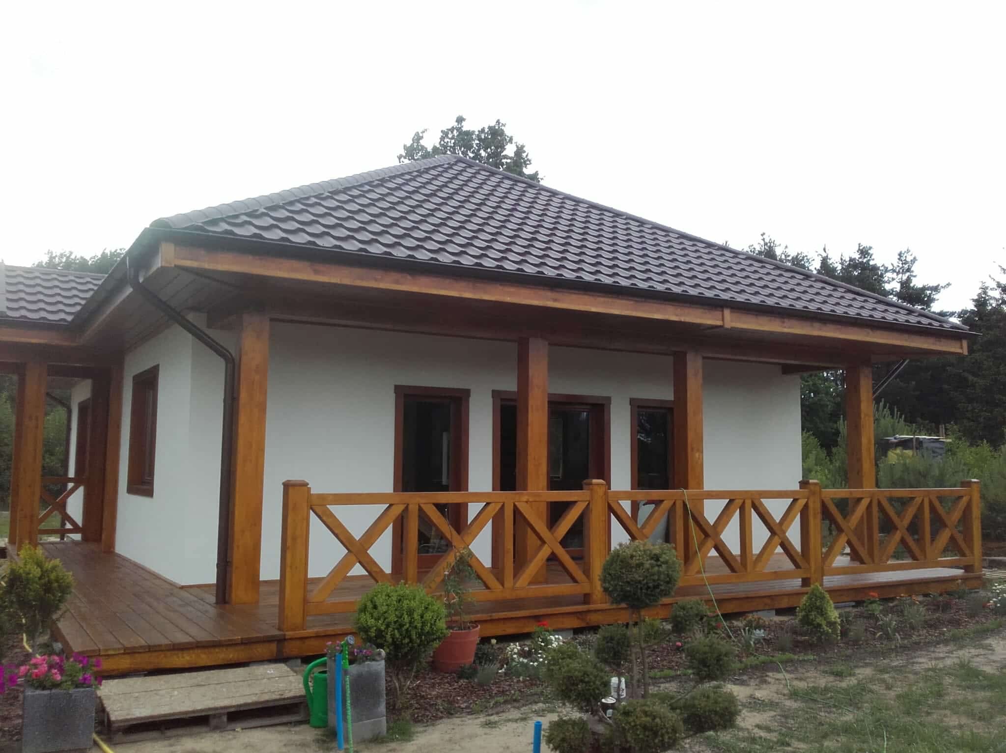 Gotowy dom szkieletowy z drewnianym tarasem i balustradą w kształcie krzyży. Brązowy dach z blachodachówki, białe ściany. Widok z zewnątrz, ogród z roślinami.