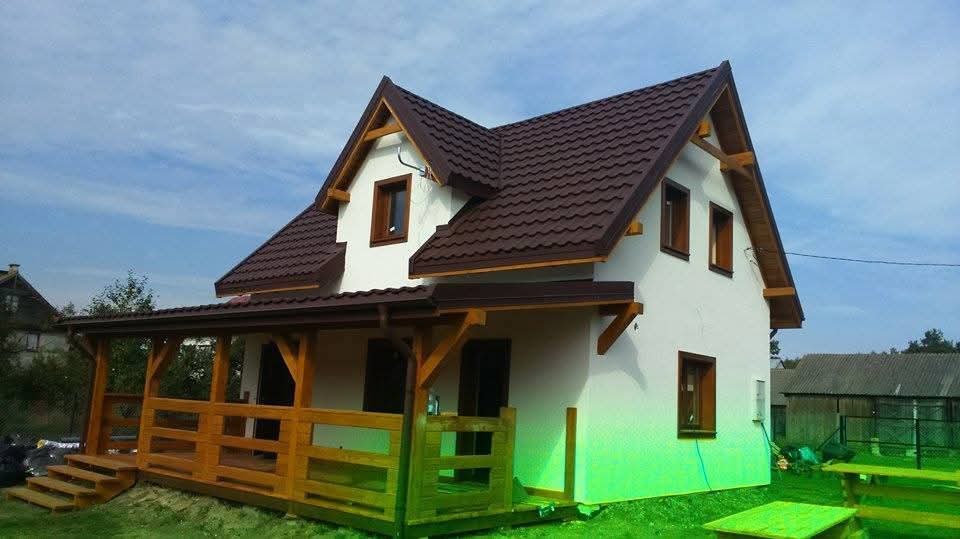 Gotowy dom szkieletowy z brązowym dachem i drewnianą werandą, widok z zewnątrz w słoneczny dzień. Charakterystyczne drewniane wykończenia okien i dachu.