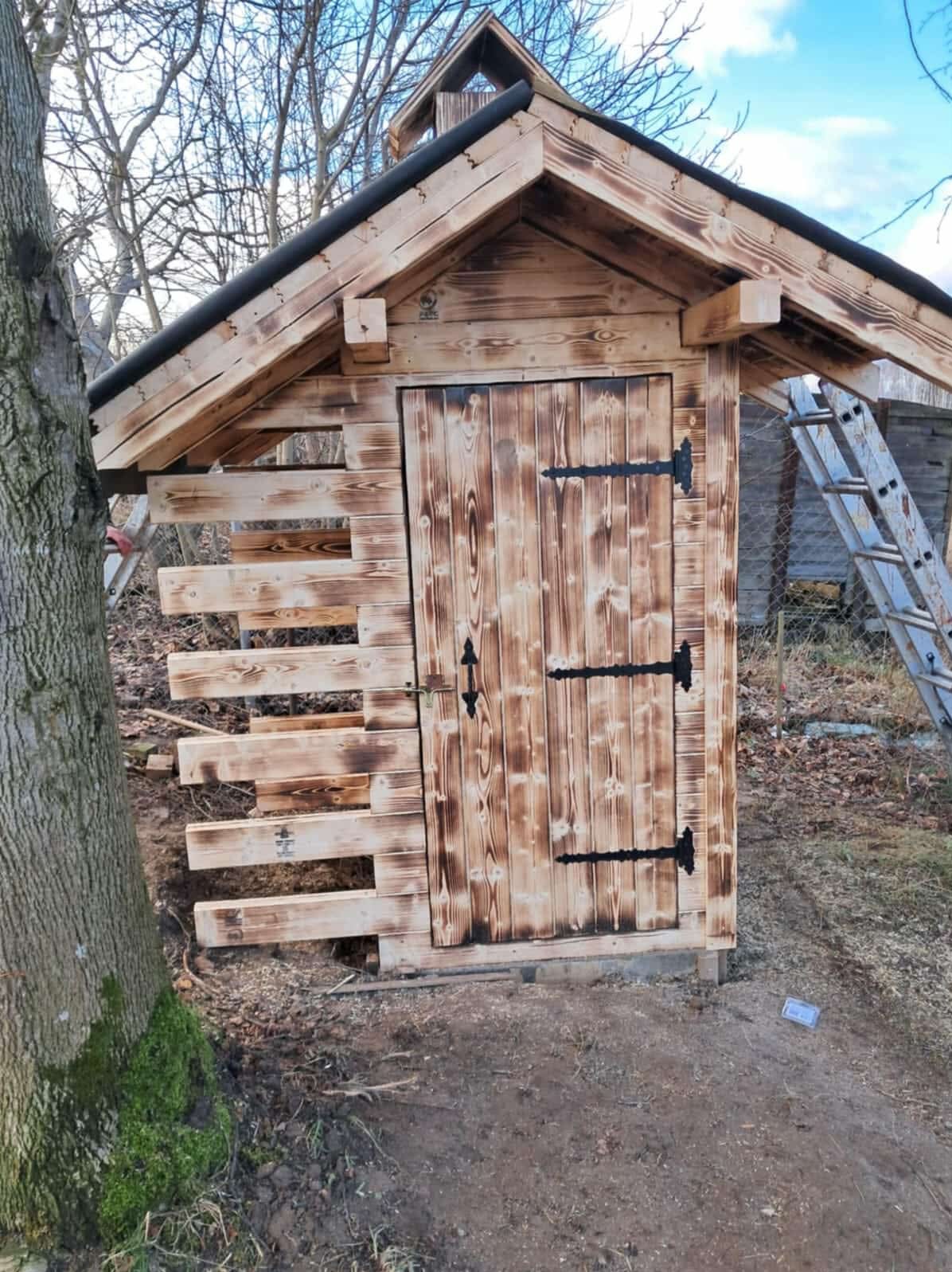 Drewniana budka z palonego drewna, czarne okucia drzwi, częściowo wykończona elewacja z desek, drabina oparta o ogrodzenie w tle, drzewo z mchem po lewej.