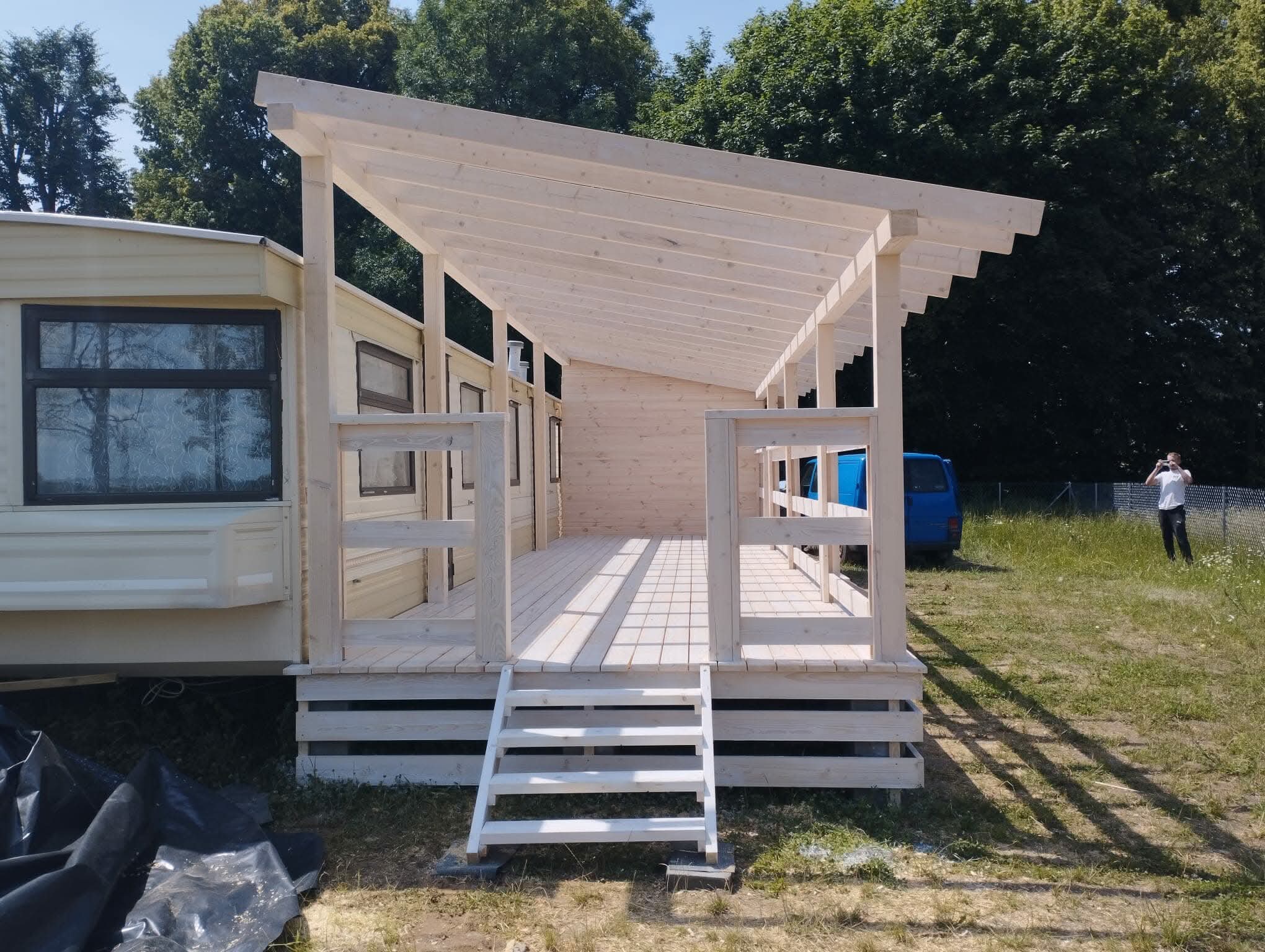 Drewniana konstrukcja z zadaszeniem, prawdopodobnie weranda lub taras, dobudowana do przyczepy kempingowej. Widoczne schody wejściowe i elementy konstrukcyjne.