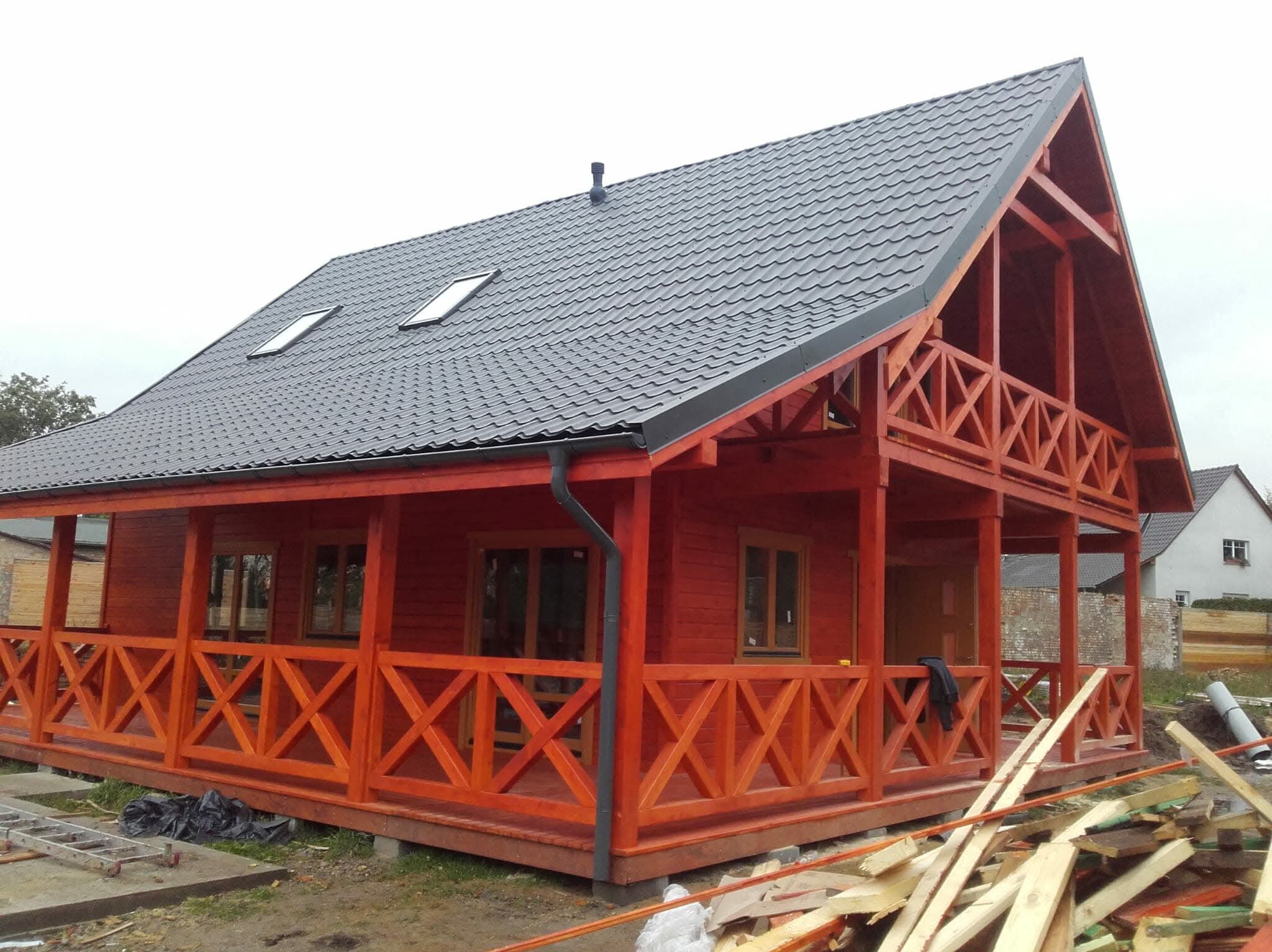 Czerwony dom szkieletowy z ciemnym dachem i drewnianą balustradą, widoczny z zewnątrz, z elementami wykończeniowymi i resztkami materiałów budowlanych wokół.