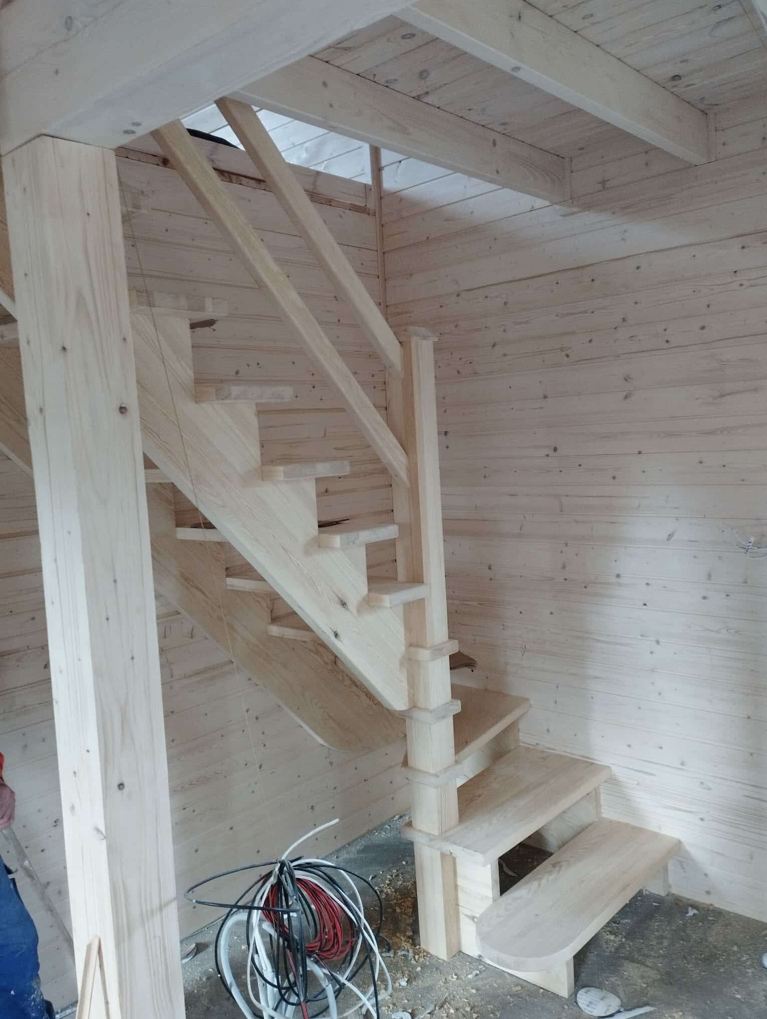 Wnętrze domu szkieletowego w trakcie budowy: drewniane schody z balustradą, ściany i sufit z desek, widoczne przewody elektryczne na podłodze.