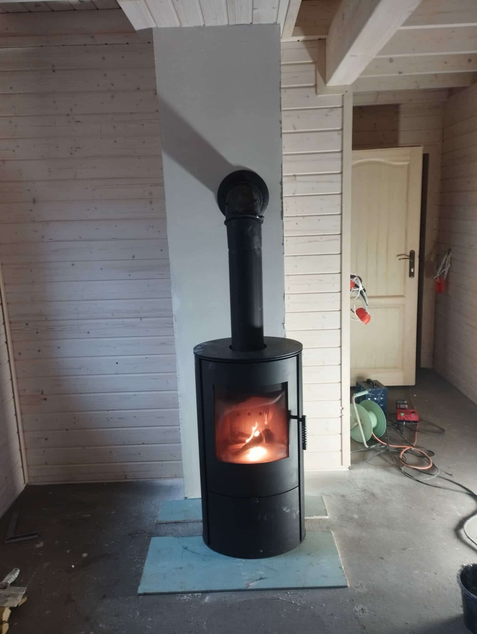 Wnętrze domu szkieletowego z czarnym piecem kominkowym na tle boazerii i białej ściany. Widoczne narzędzia i kable sugerują trwające prace wykończeniowe.