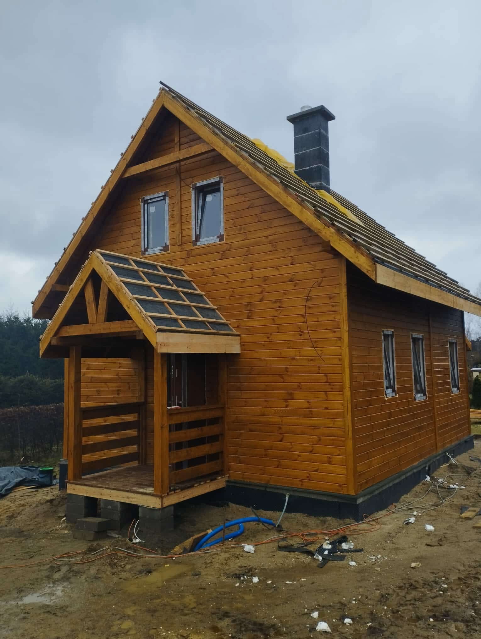 Drewniany dom szkieletowy w trakcie budowy, widoczne elementy konstrukcyjne dachu i komin. Dom z gankiem, na fundamencie, otoczony ziemią i przewodami.