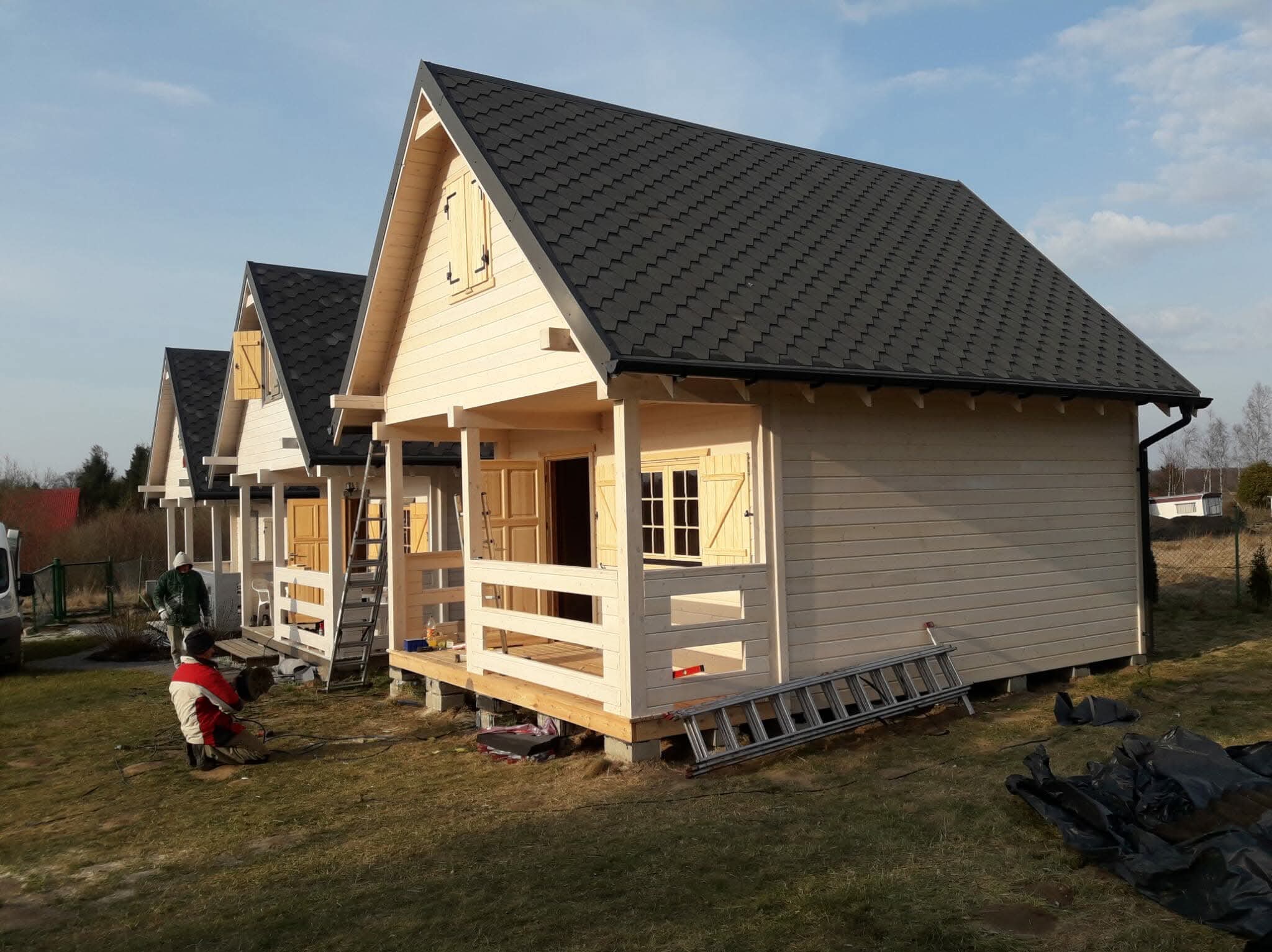 Trzy identyczne, jasne domki szkieletowe z ciemnymi dachami, ustawione w rzędzie. Na pierwszym planie pracownik w trakcie wykonywania prac wykończeniowych.