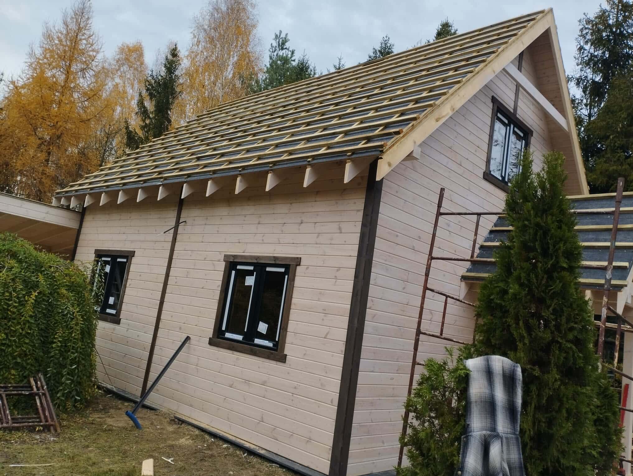 Drewniany dom w trakcie budowy dachu, widoczne łaty dachowe. Ściany w jasnym odcieniu, okna z ciemnymi ramami. Na zewnątrz drabina i zieleń.