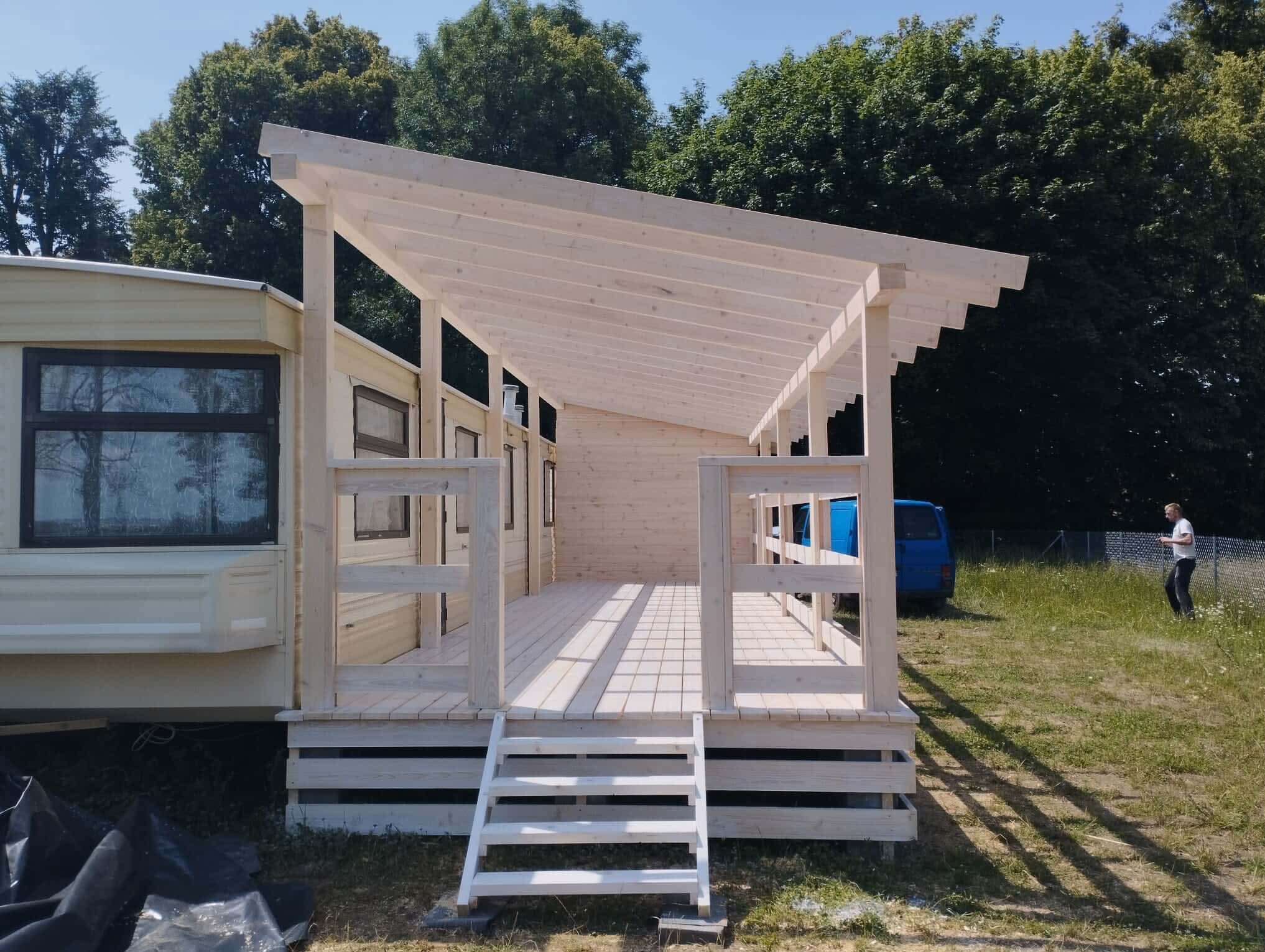 Drewniana dobudówka z zadaszeniem przyczepy kempingowej, jasne drewno, schody wejściowe, widok z zewnątrz w słoneczny dzień, drzewa w tle.
