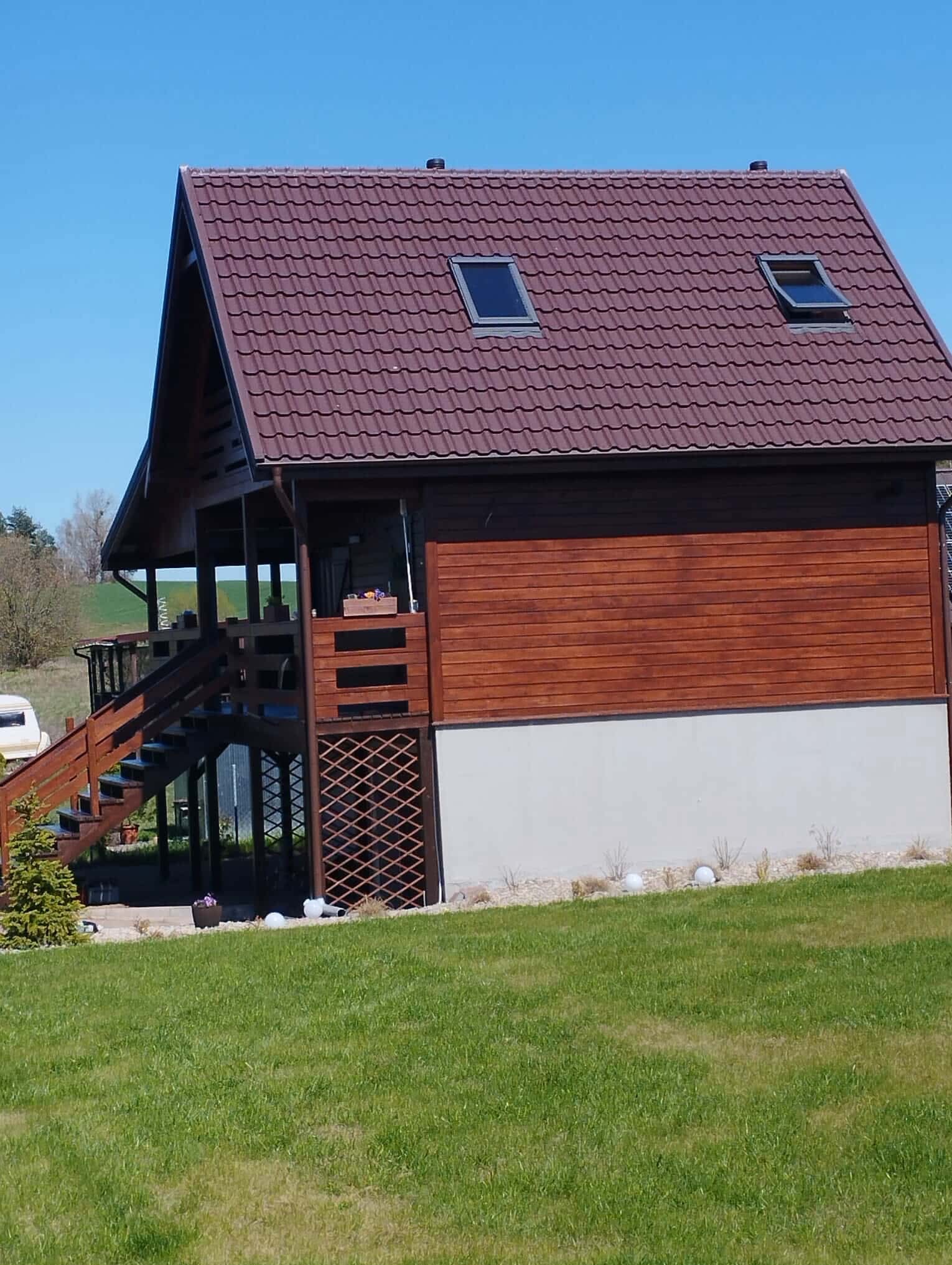 Gotowy dom szkieletowy z brązową dachówką i drewnianą elewacją, widoczny z poziomu trawnika w Ostródzie. Uwagę zwracają okna dachowe i zadaszony taras.