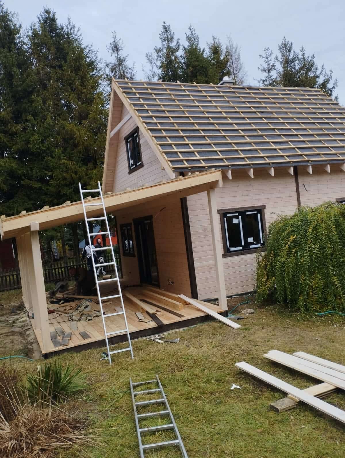 Wznoszenie domu szkieletowego: drewniana konstrukcja z częściowo ułożonym dachem, drabina oparta o budynek, widoczne elementy wykończeniowe i narzędzia na trawie.
