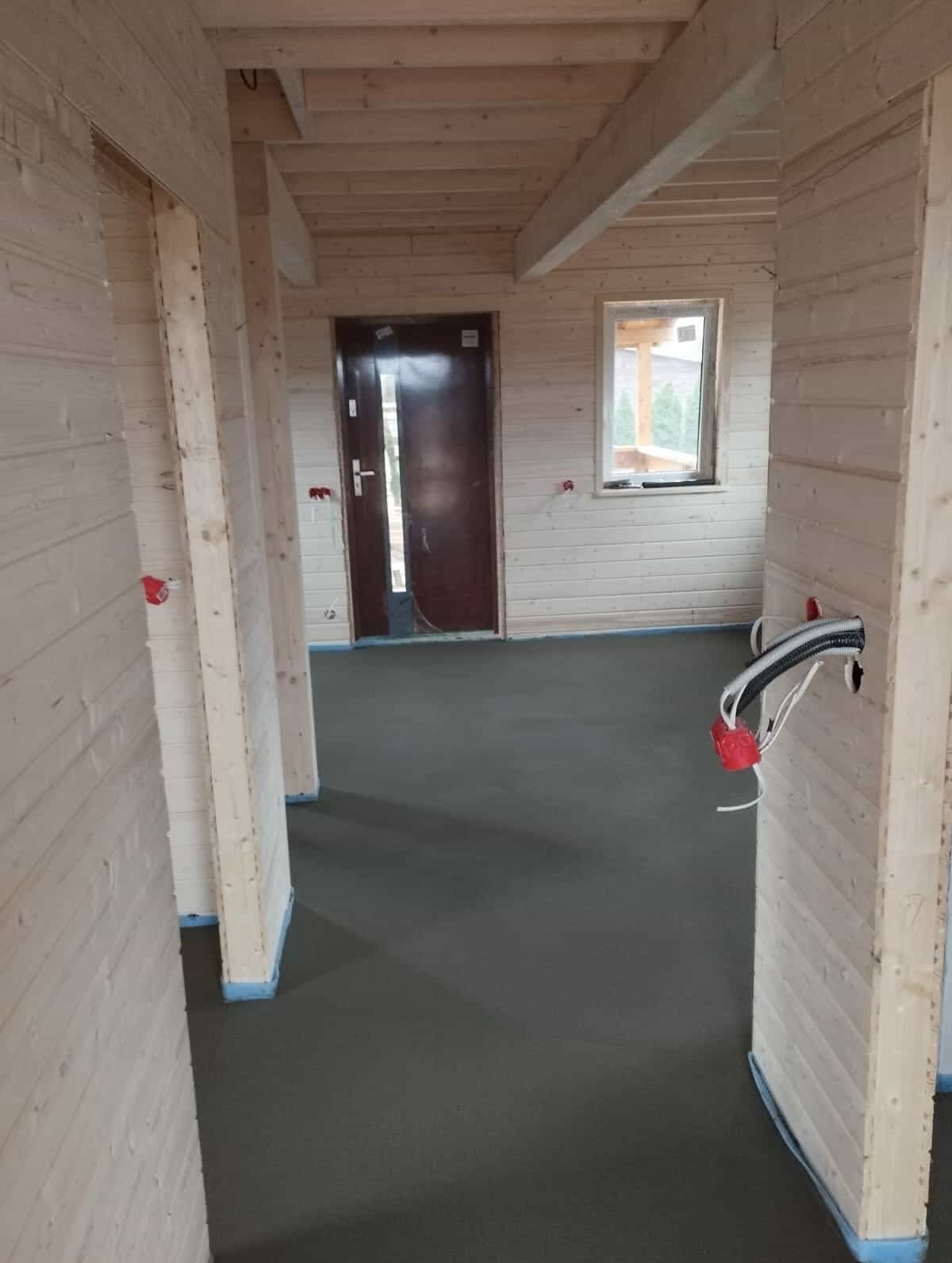 Wnętrze domu szkieletowego w trakcie budowy: surowe drewniane ściany, wylana posadzka, wystające przewody elektryczne. Widoczne okno i drzwi wejściowe.