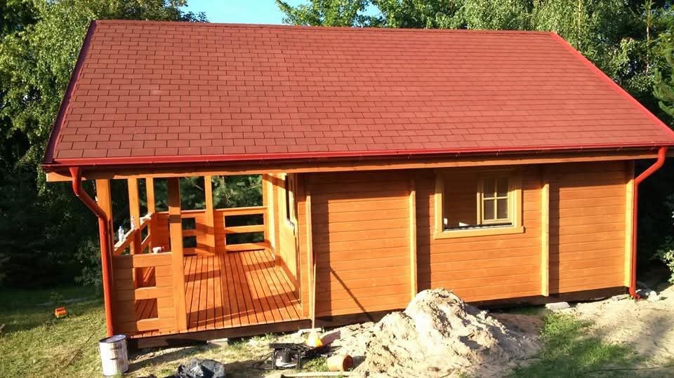 Drewniany domek letniskowy z czerwonym dachem, widoczny z zewnątrz, z elementami wykończeniowymi i stertą ziemi obok, na tle zieleni.