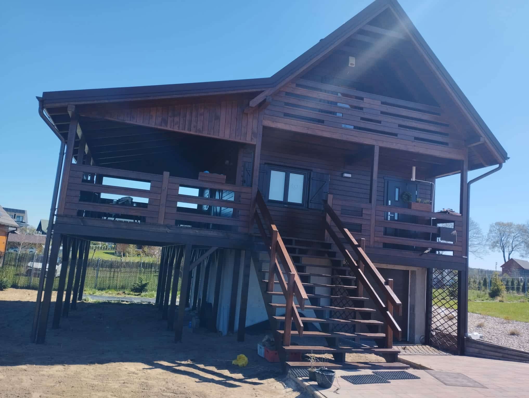 Drewniany dom szkieletowy na palach z tarasem i schodami, wykończony ciemnym drewnem, na tle błękitnego nieba i zieleni. Nietypowa konstrukcja.