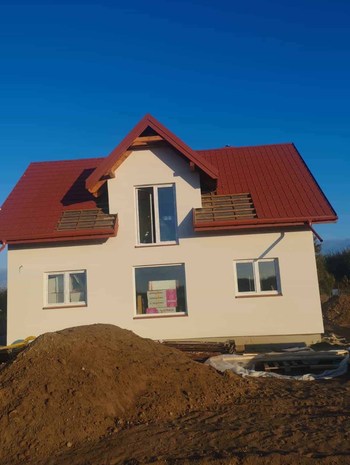 Dwukondygnacyjny dom z czerwonym dachem w trakcie budowy, widoczne okna i sterta ziemi na pierwszym planie. Częściowo zamontowane elementy balkonu.