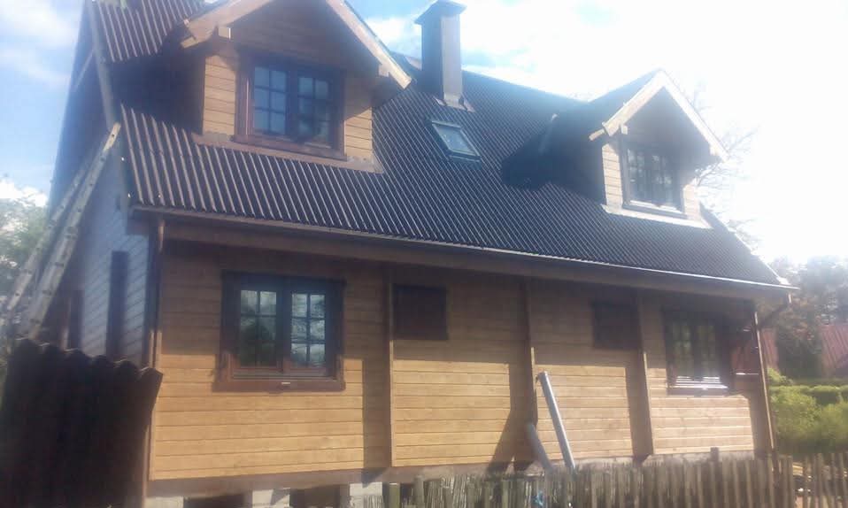 Gotowy dom szkieletowy z ciemnym dachem, widoczna drabina oparta o ścianę. Drewniana elewacja w ciepłym odcieniu brązu, okna z ciemnymi ramami.