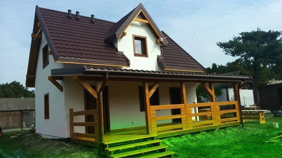 Gotowy dom szkieletowy z brązowym dachem i drewnianą werandą, otoczony zielonym trawnikiem. Widoczne detale wykończenia i elementy konstrukcyjne.