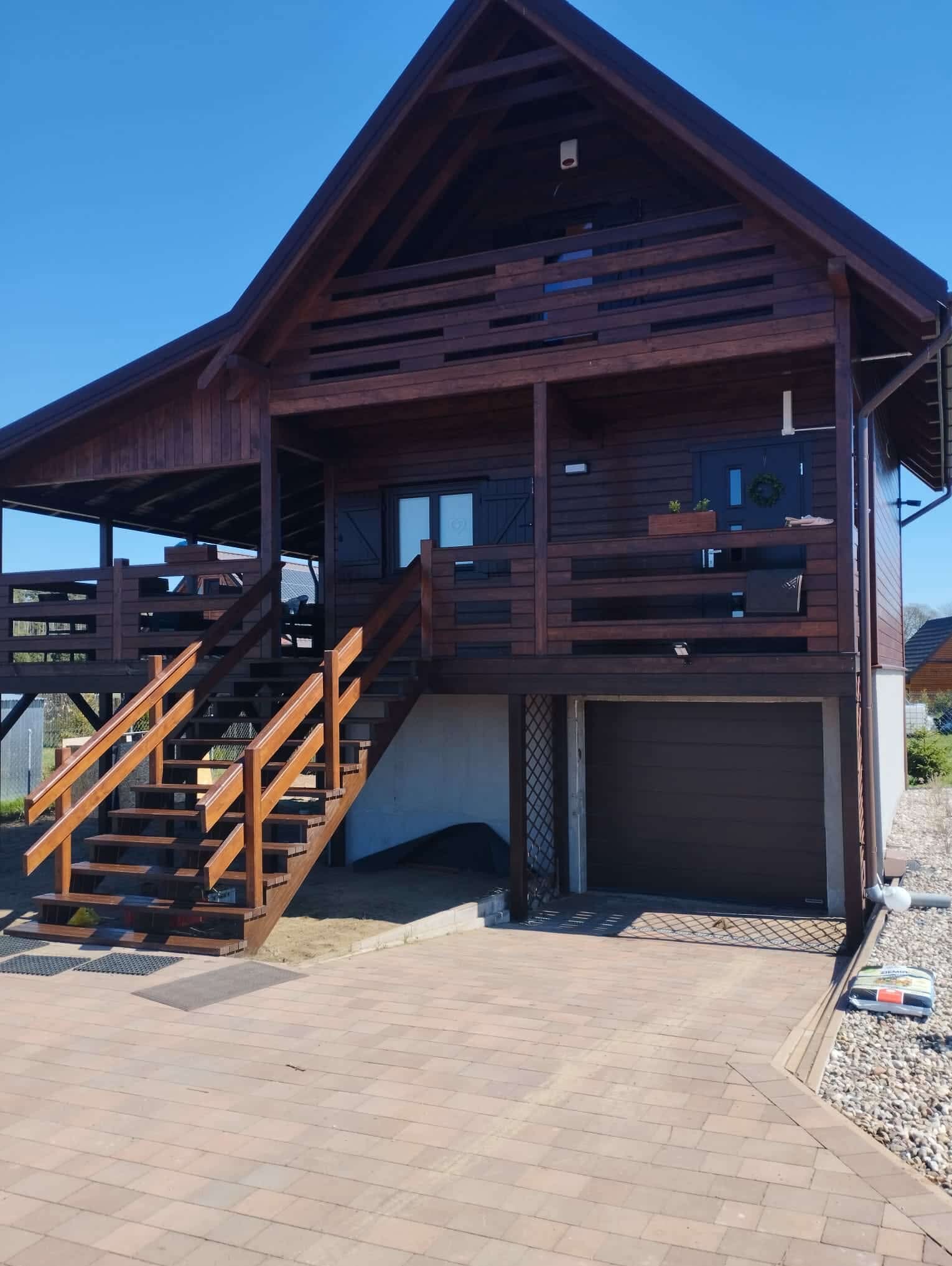 Dwukondygnacyjny dom szkieletowy z ciemnego drewna z balkonem i schodami zewnętrznymi, garaż w bryle budynku, kostka brukowa na podjeździe, błękitne niebo.