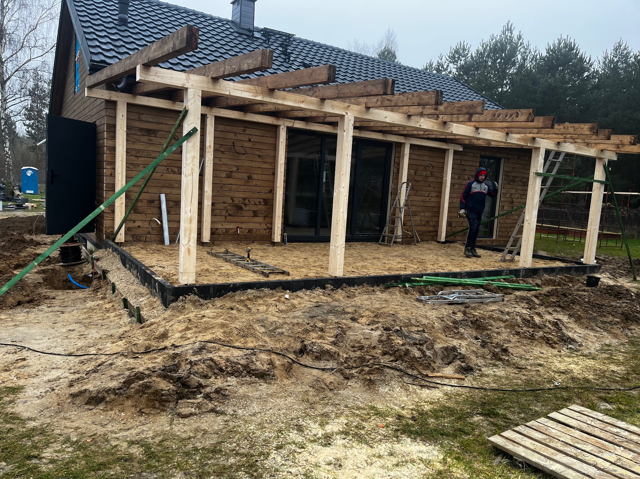 Budowa drewnianej pergoli przy domu z ciemną dachówką. Widoczne elementy konstrukcyjne, teren przygotowany pod wylewkę. Pracownik w tle.