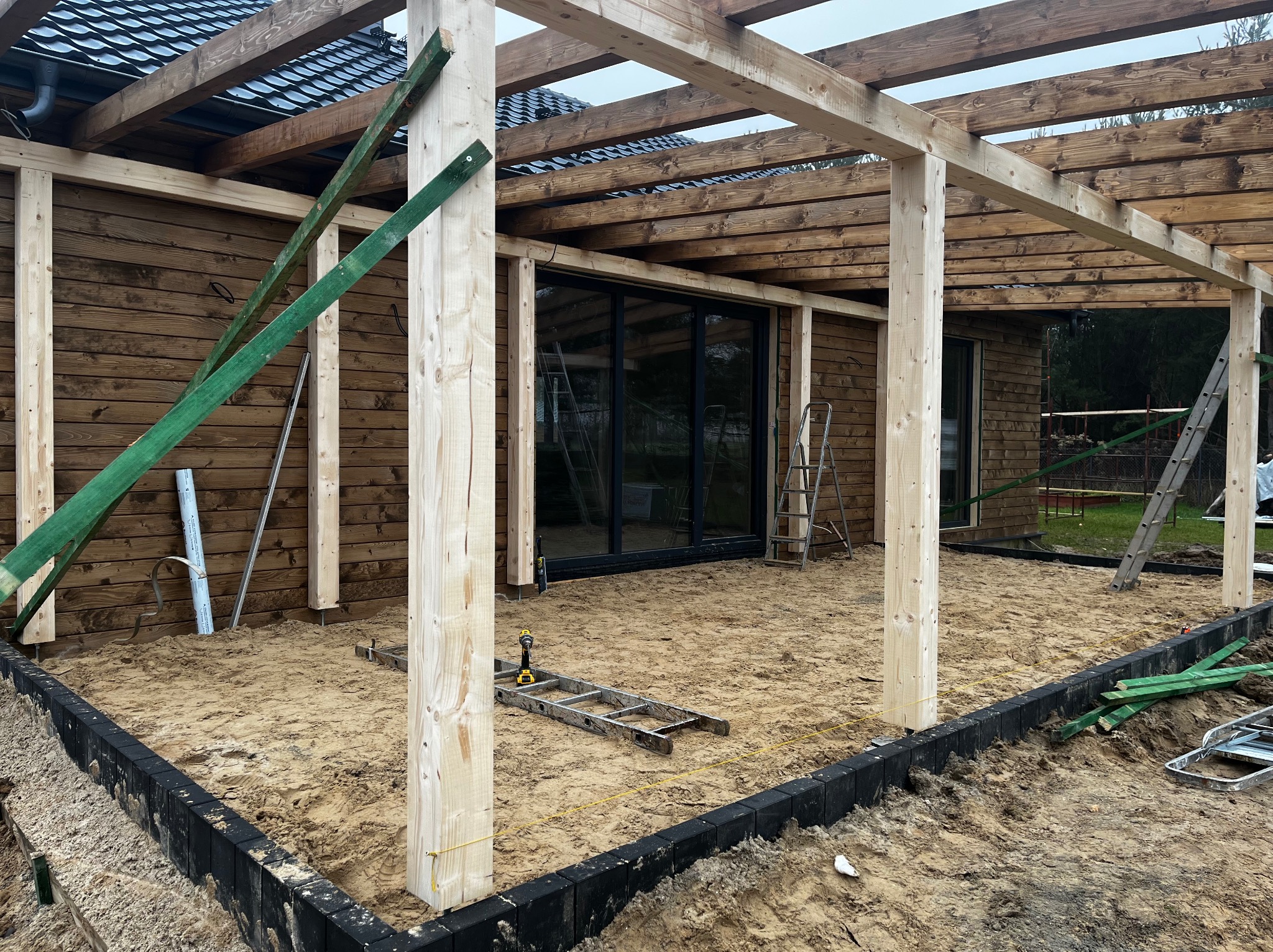 Drewniana konstrukcja zadaszenia tarasu w trakcie budowy, widoczne słupy, belki, podłoże z piasku i obrzeża z kostki. W tle ściana budynku z oknami.