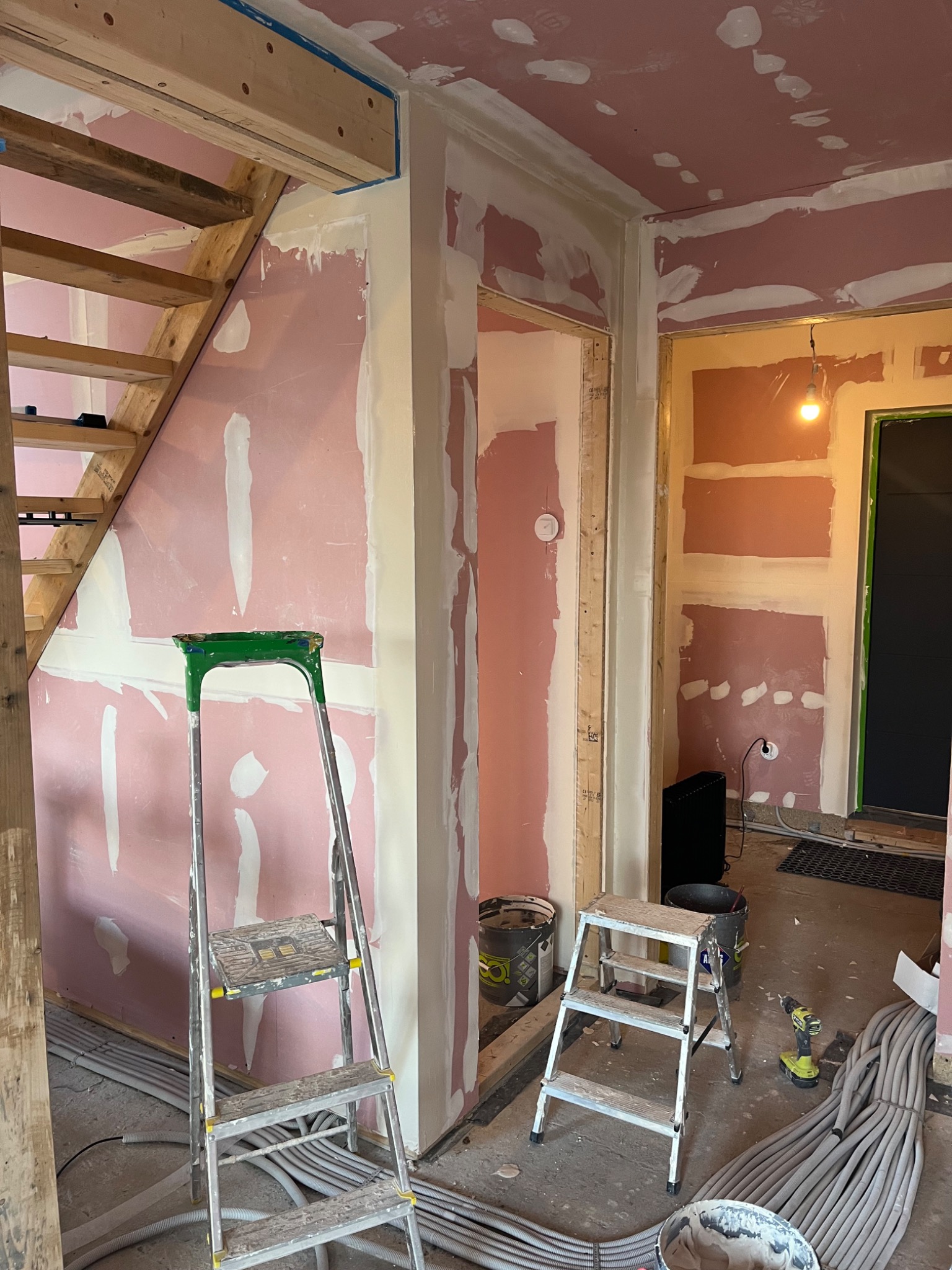 Wnętrze w trakcie prac wykończeniowych: ściany z płyt gipsowo-kartonowych, częściowo pokryte gładzią, drabiny, przewody elektryczne. Widoczne elementy drewniane schodów.