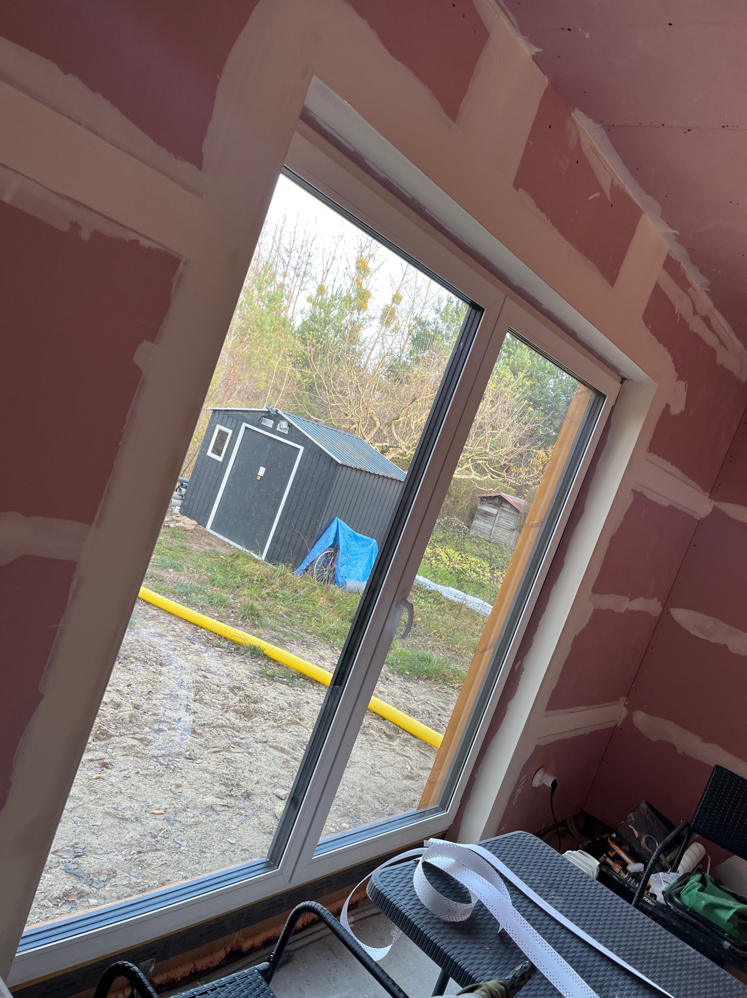 Poddasze z oknem dachowym w trakcie prac wykończeniowych. Widoczne płyty kartonowo-gipsowe i szpachlowane łączenia. Na zewnątrz widok na ogród z domkiem narzędziowym.