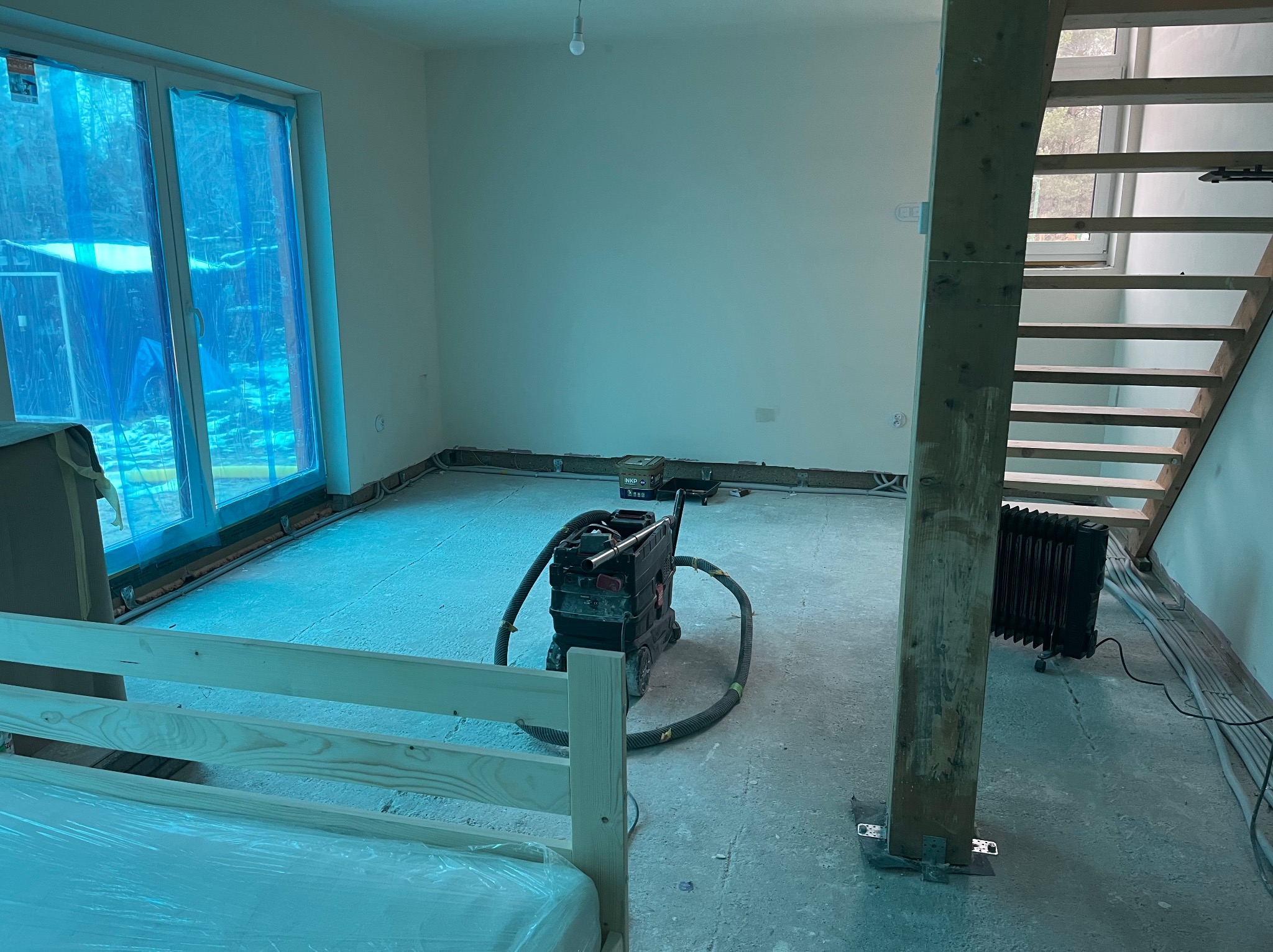 Wnętrze w trakcie remontu: zabezpieczone okno folią, szlifierka do gładzi na posadzce, drewniane schody, grzejnik elektryczny, widoczne kable i instalacje.