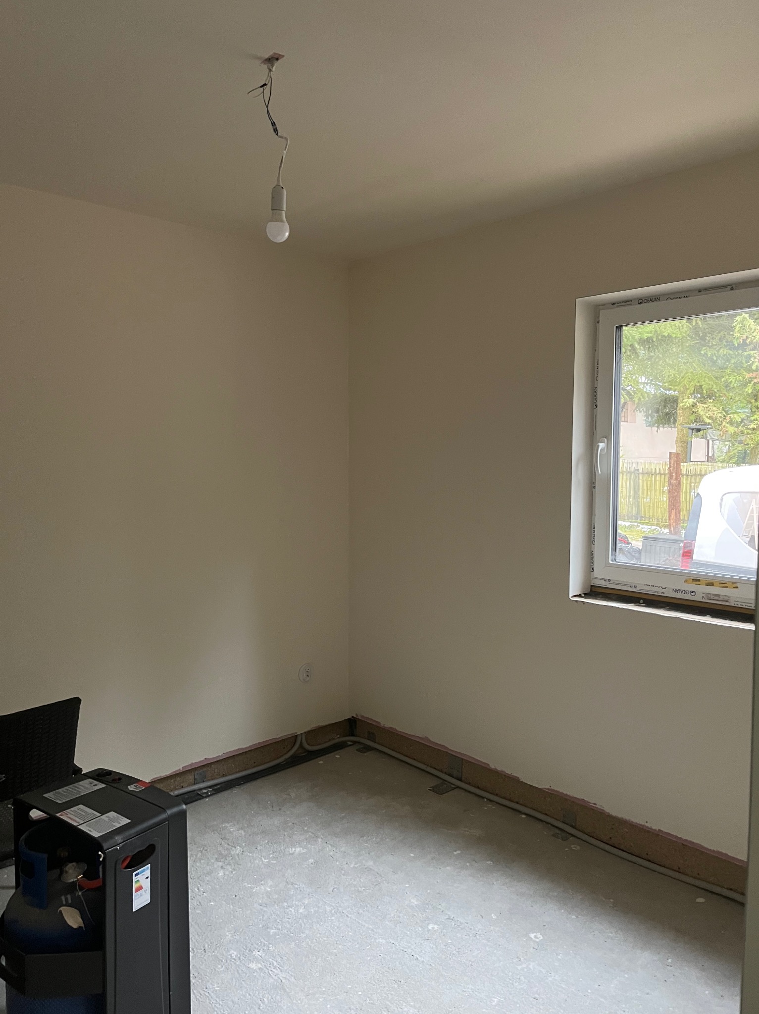 Surowe wnętrze pokoju z oknem, przygotowane do prac wykończeniowych. Widoczne ściany w jasnym odcieniu, surowa podłoga i prowizoryczne oświetlenie.