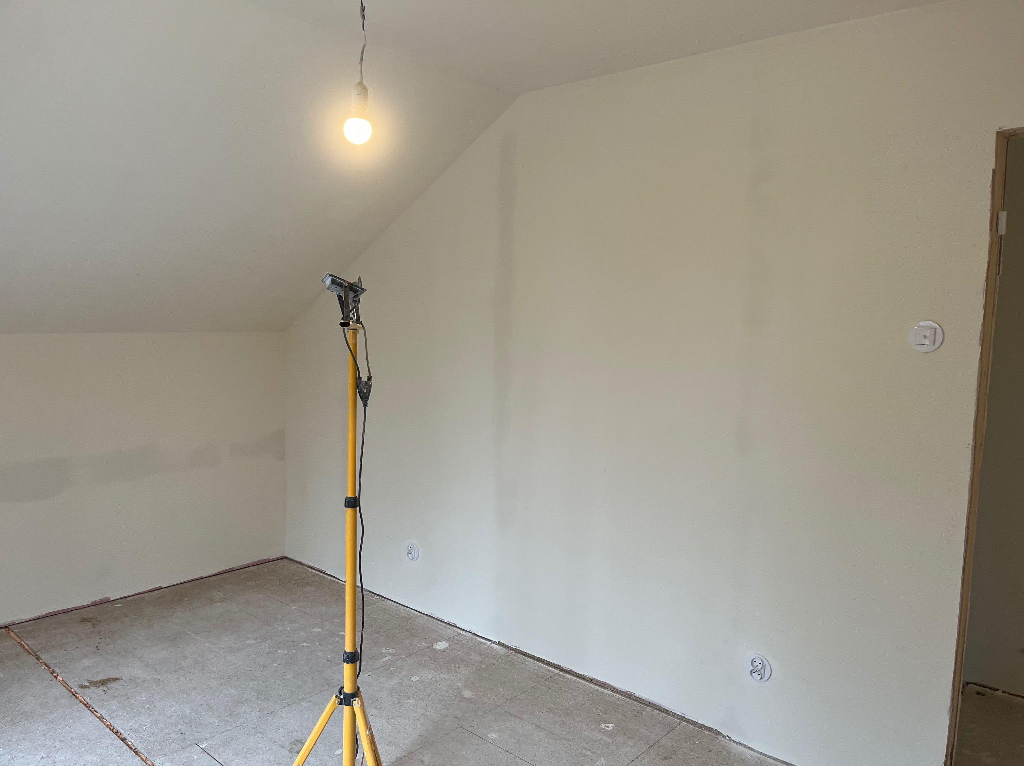Poddasze po malowaniu na jasny kolor, widoczna lampa na statywie, gniazdka elektryczne i fragment podłogi. Świeżo pomalowane ściany w pokoju na poddaszu.