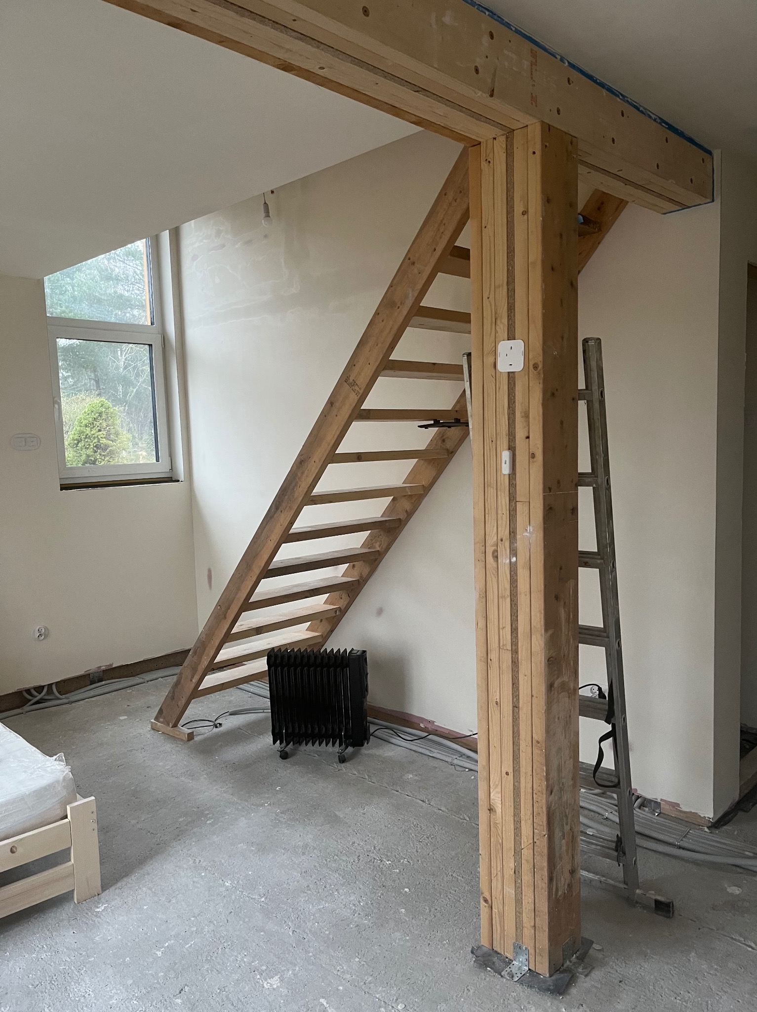 Surowe drewniane schody w jasnym wnętrzu w trakcie prac wykończeniowych. Widoczne kable, grzejnik i drabina. Elementy konstrukcyjne z drewna.