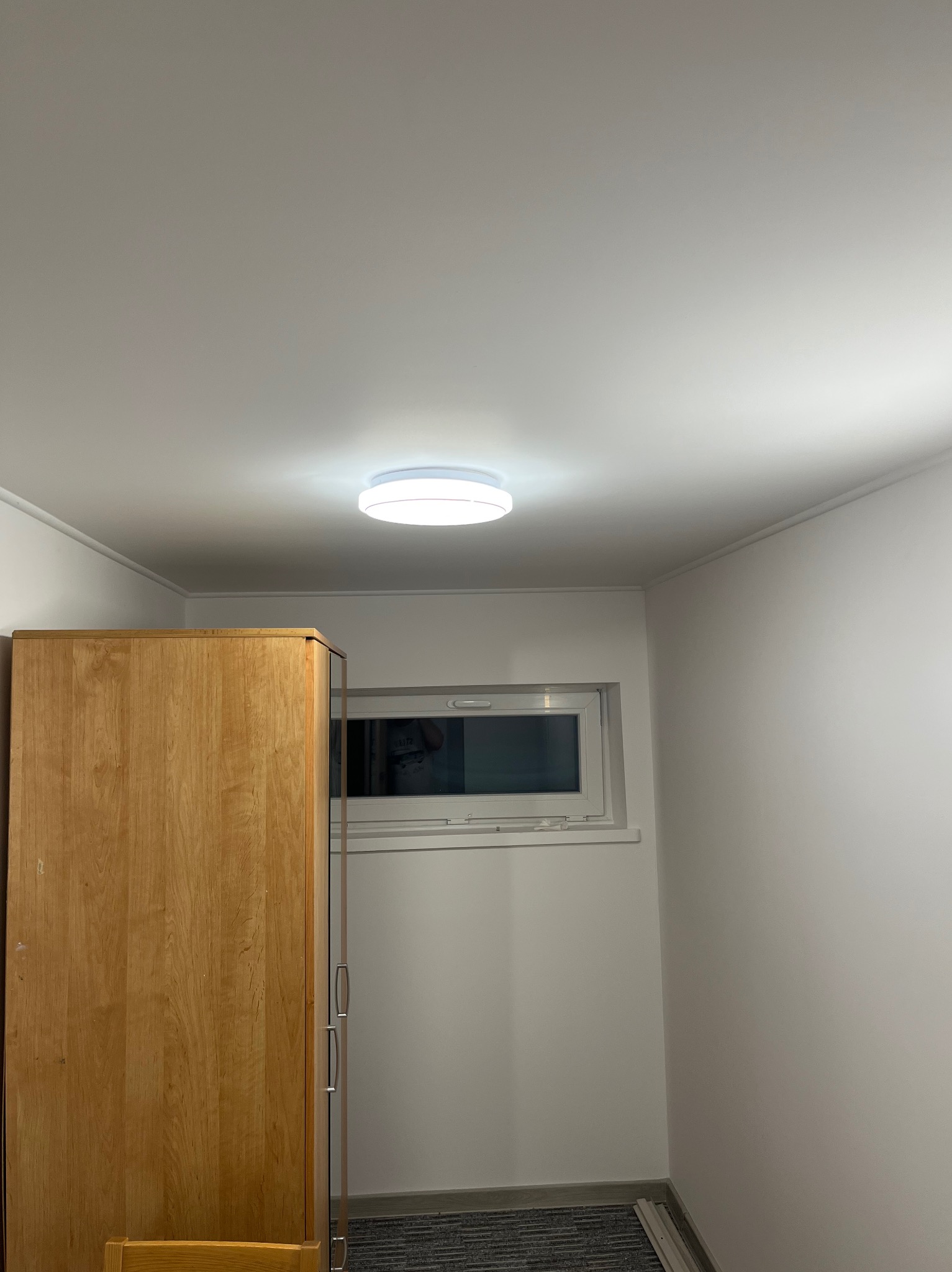 Wnętrze pokoju po remoncie: gładkie, białe ściany, okno z białymi ramami, drewniana szafa i nowoczesna lampa sufitowa. Minimalistyczny design i świeżo odmalowane powierzchnie.