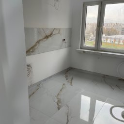 Usługi glazurnicze Piotr Pisarczyk - Jasne wnętrze po remoncie: marmurowe płytki na podłodze i ścianie, okno z widokiem na miasto. Widoczne kable elektryczne, sugerujące prace wykończeniowe w toku.