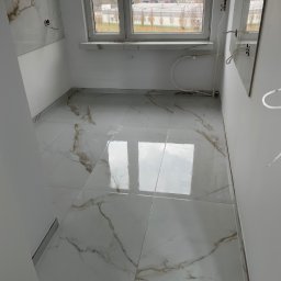 Usługi glazurnicze Piotr Pisarczyk - Jasne, marmurowe płytki podłogowe w wykończonym wnętrzu łazienki z oknem. Widoczne fragmenty instalacji i gniazdek elektrycznych.