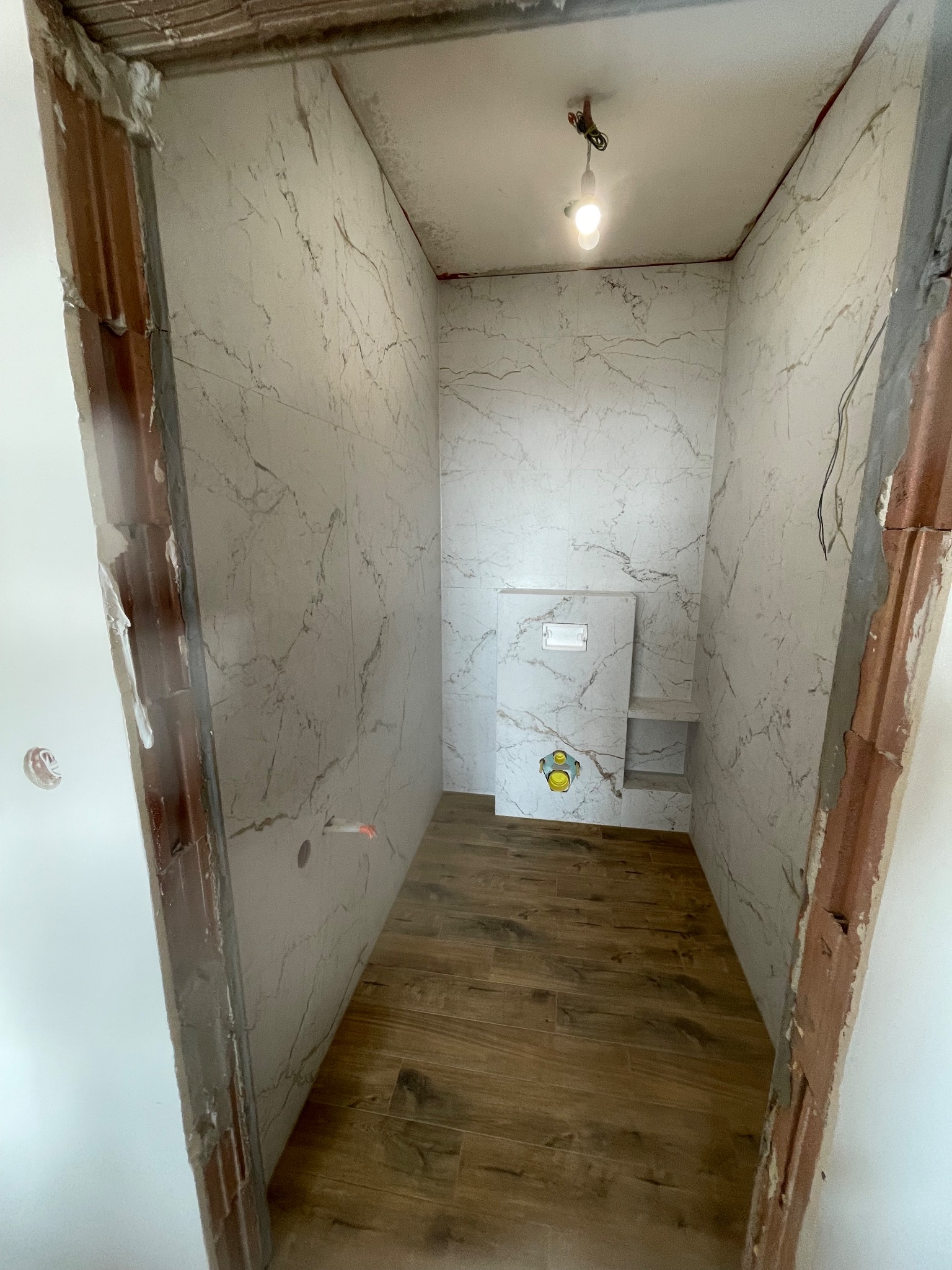 Wnętrze małej łazienki w trakcie remontu: marmurowe płytki na ścianach, drewnopodobna podłoga, zabudowana toaleta. Widoczne surowe wykończenie ościeżnicy.