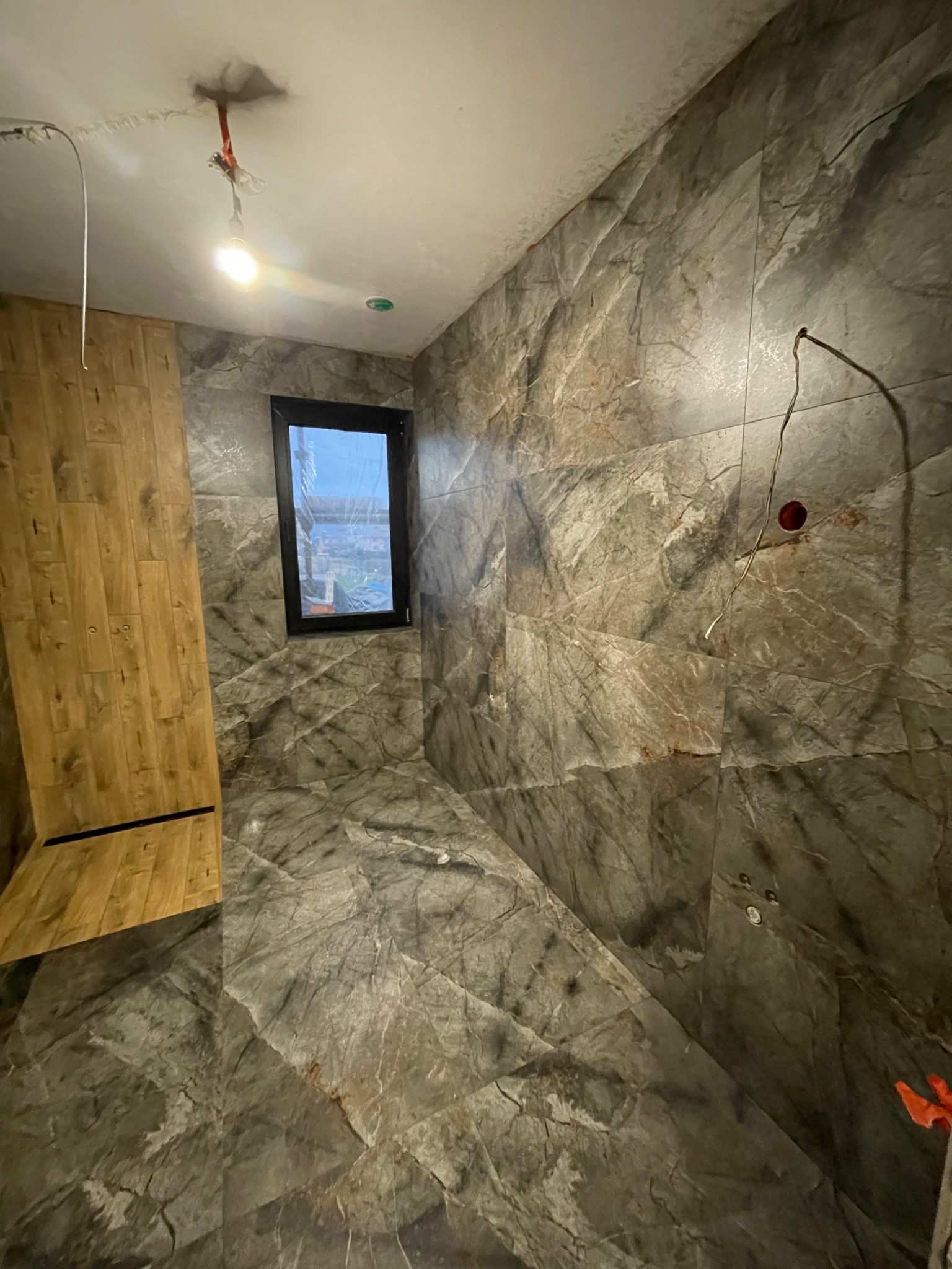 Wnętrze łazienki w trakcie wykańczania: marmurowe płytki na ścianach i podłodze, drewniana boazeria, okno z czarną ramą, widoczne przewody elektryczne.