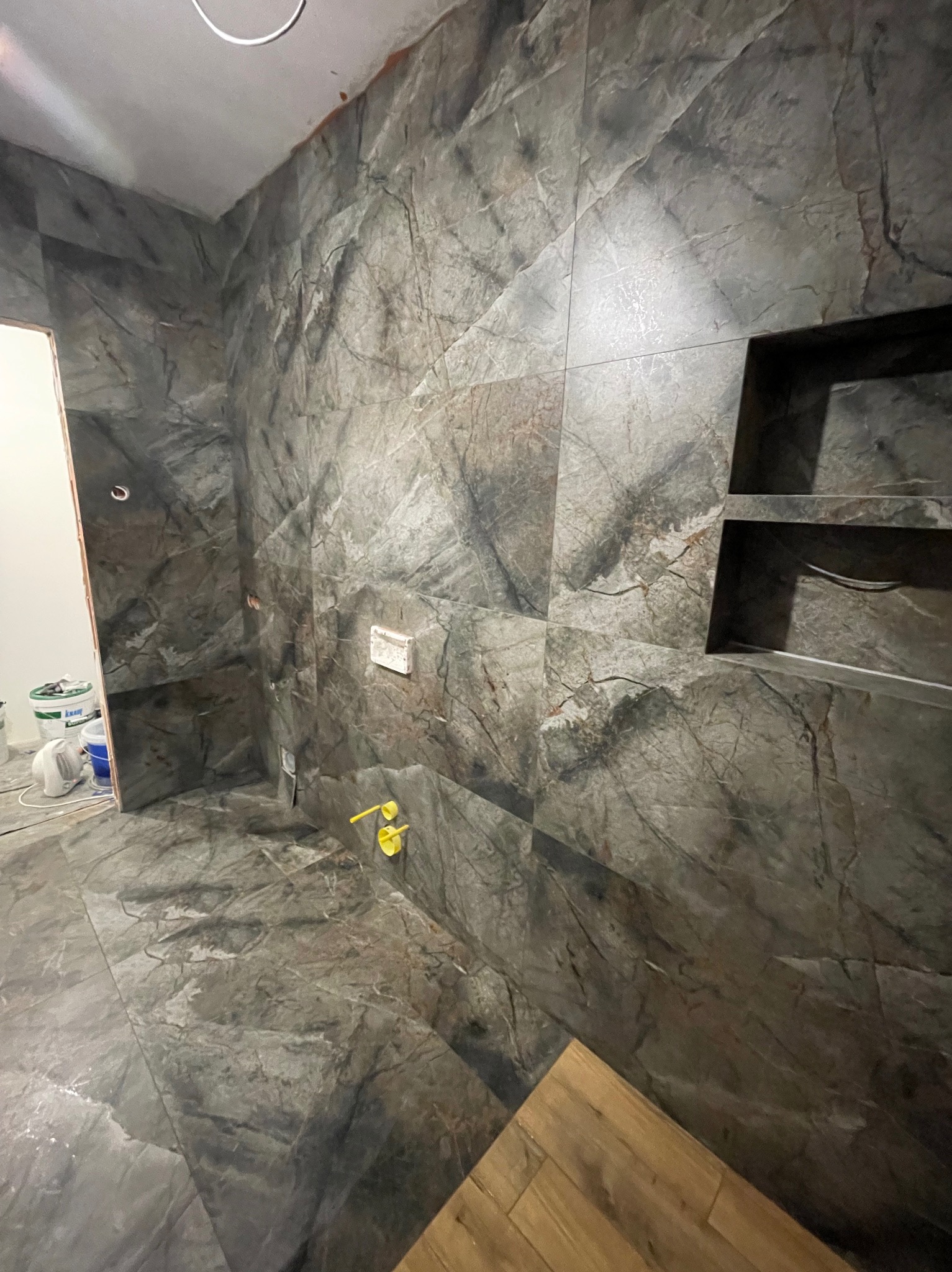 Wykończona łazienka z szarymi płytkami imitującymi marmur, wnęka w ścianie i żółte elementy instalacji. Widoczne wiaderka z farbą.