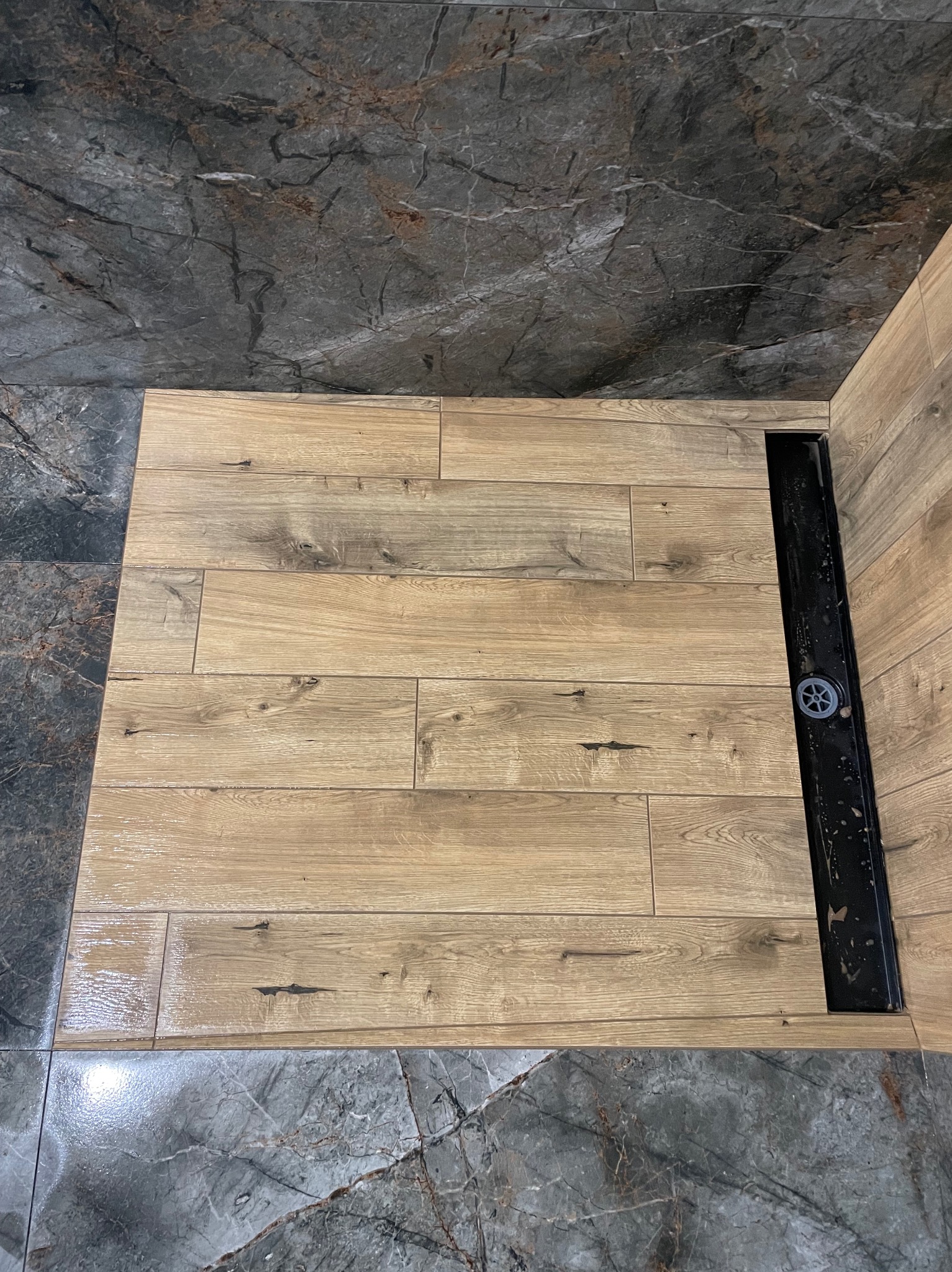 Ułożone płytki imitujące drewno w kabinie prysznicowej z odpływem liniowym, kontrastujące z szarymi płytkami imitującymi marmur na ścianach i podłodze.