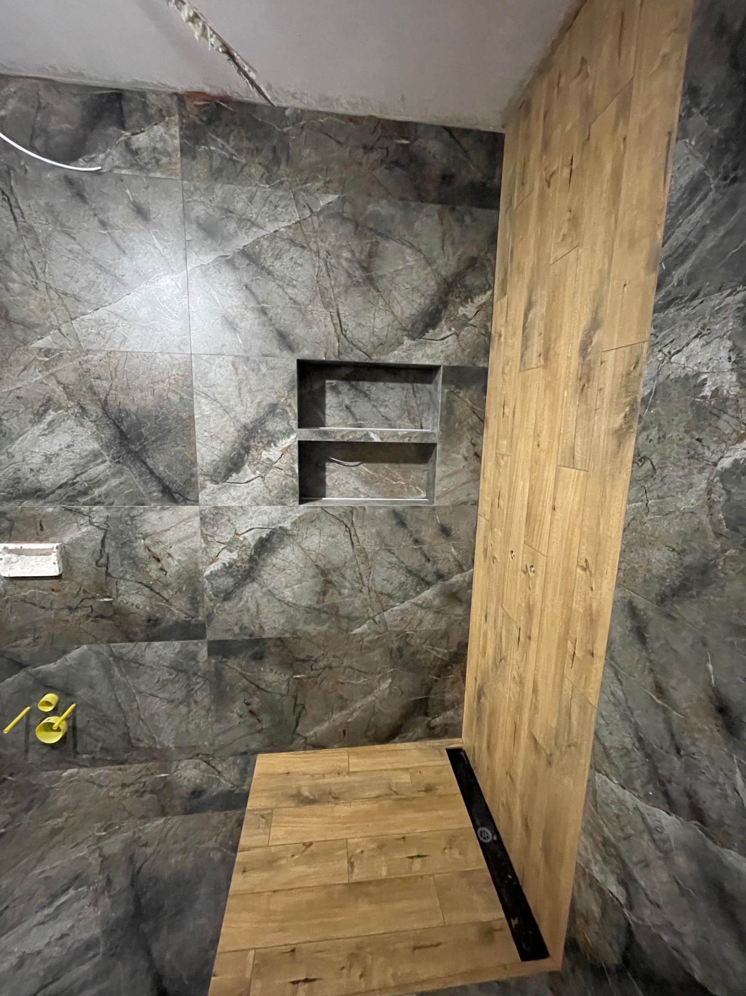 Wykończona łazienka z szarymi płytkami imitującymi marmur i drewnopodobnymi panelami. W ścianie wnęka z półkami. Widoczny odpływ liniowy.