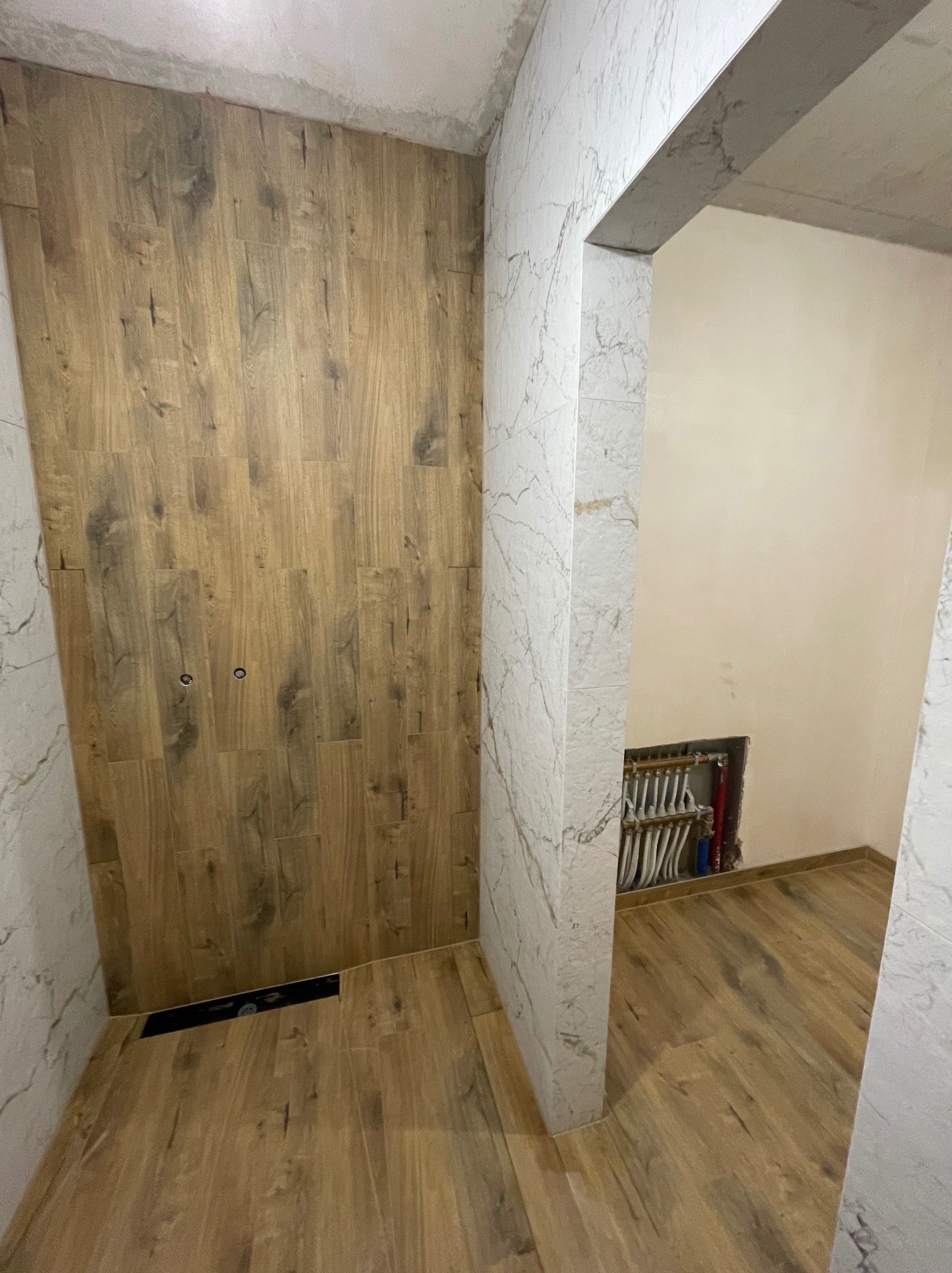 Wykończona łazienka z drewnopodobnymi płytkami na ścianie i podłodze, marmurowe płytki na ścianach, widoczna instalacja wodna w ścianie.