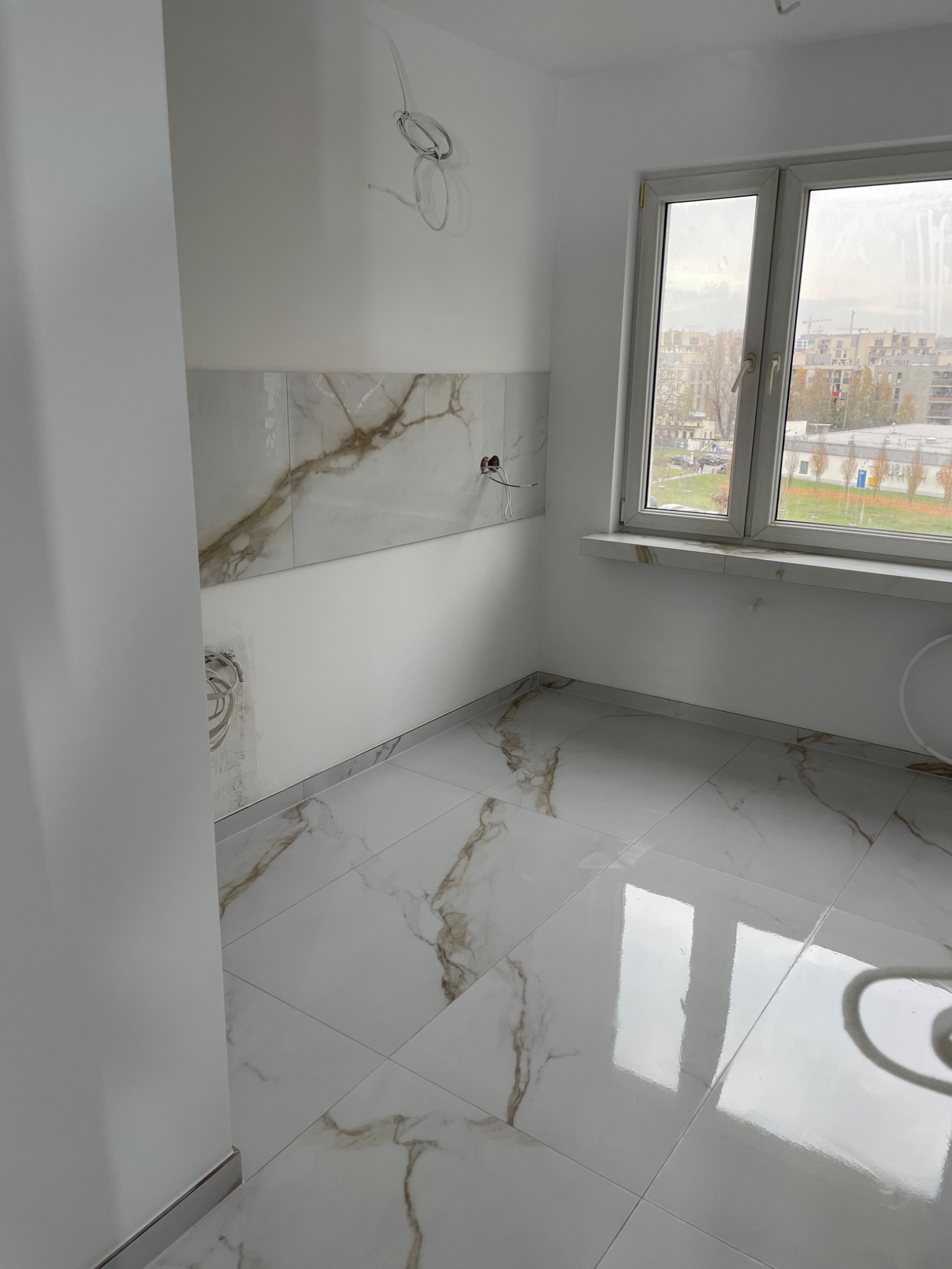 Jasne wnętrze po remoncie: marmurowe płytki na podłodze i ścianie, okno z widokiem na miasto. Widoczne kable elektryczne, sugerujące prace wykończeniowe w toku.