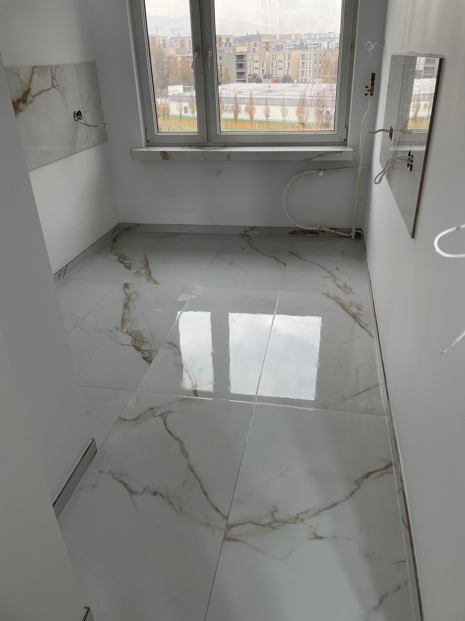 Jasne, marmurowe płytki podłogowe w wykończonym wnętrzu łazienki z oknem. Widoczne fragmenty instalacji i gniazdek elektrycznych.