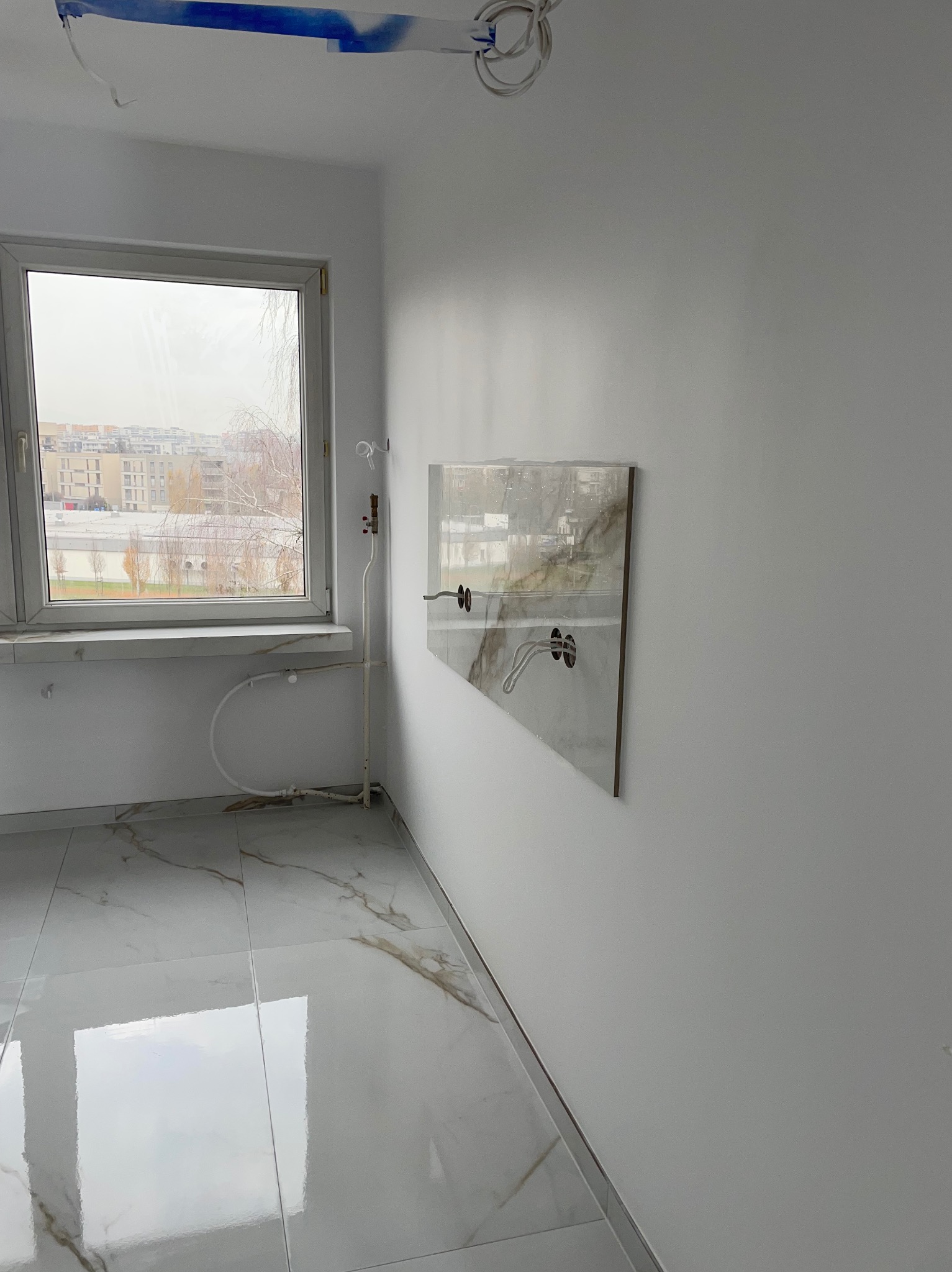 Białe wnętrze z marmurową podłogą i płytką na ścianie, przygotowane pod montaż armatury. Widoczne instalacje wodne i elektryczne. Okno z widokiem na miasto.