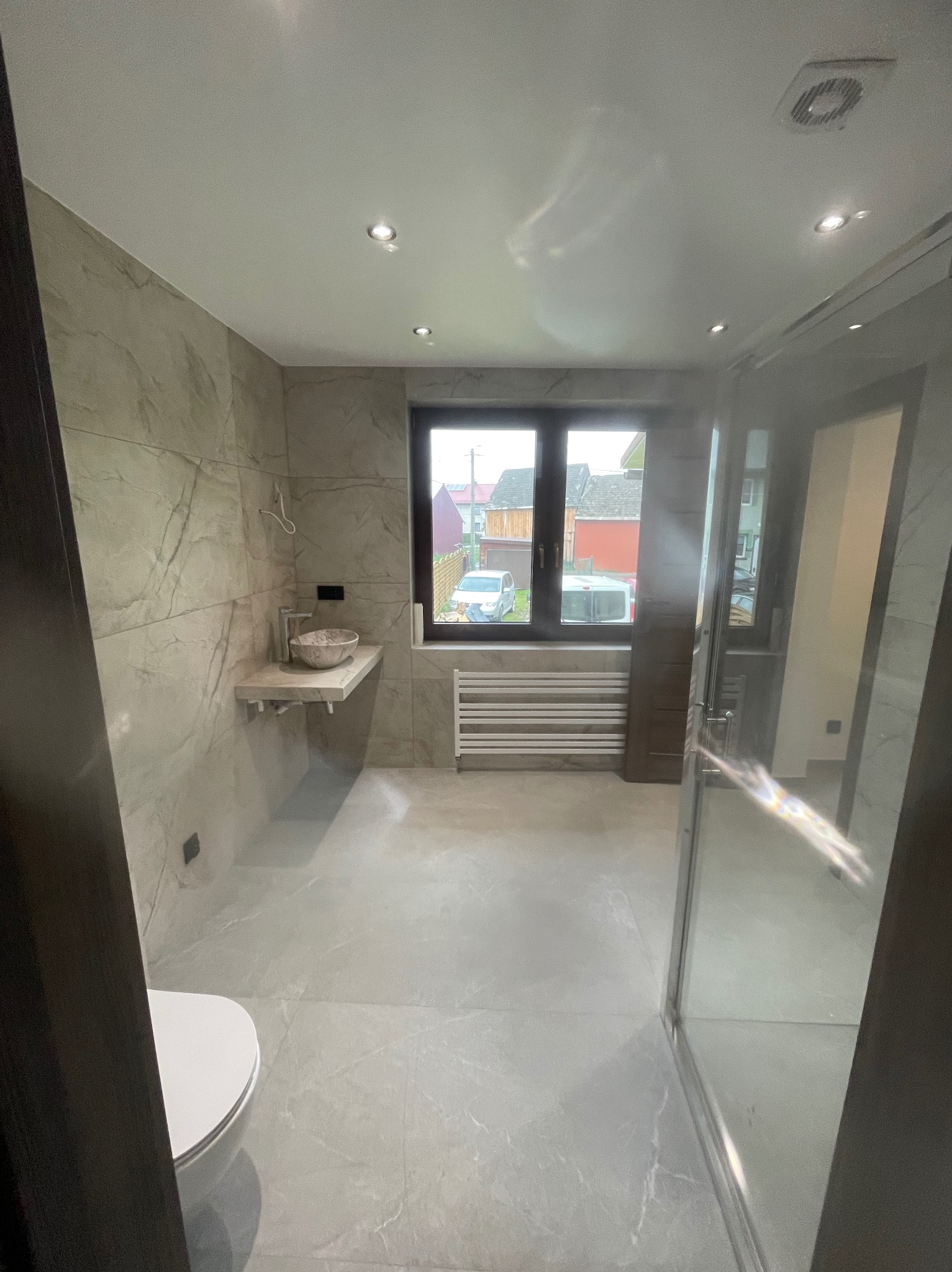 Nowoczesna łazienka z marmurowymi płytkami, wiszącą umywalką i grzejnikiem pod oknem. Kabina prysznicowa ze szklanymi drzwiami. Minimalistyczny design.