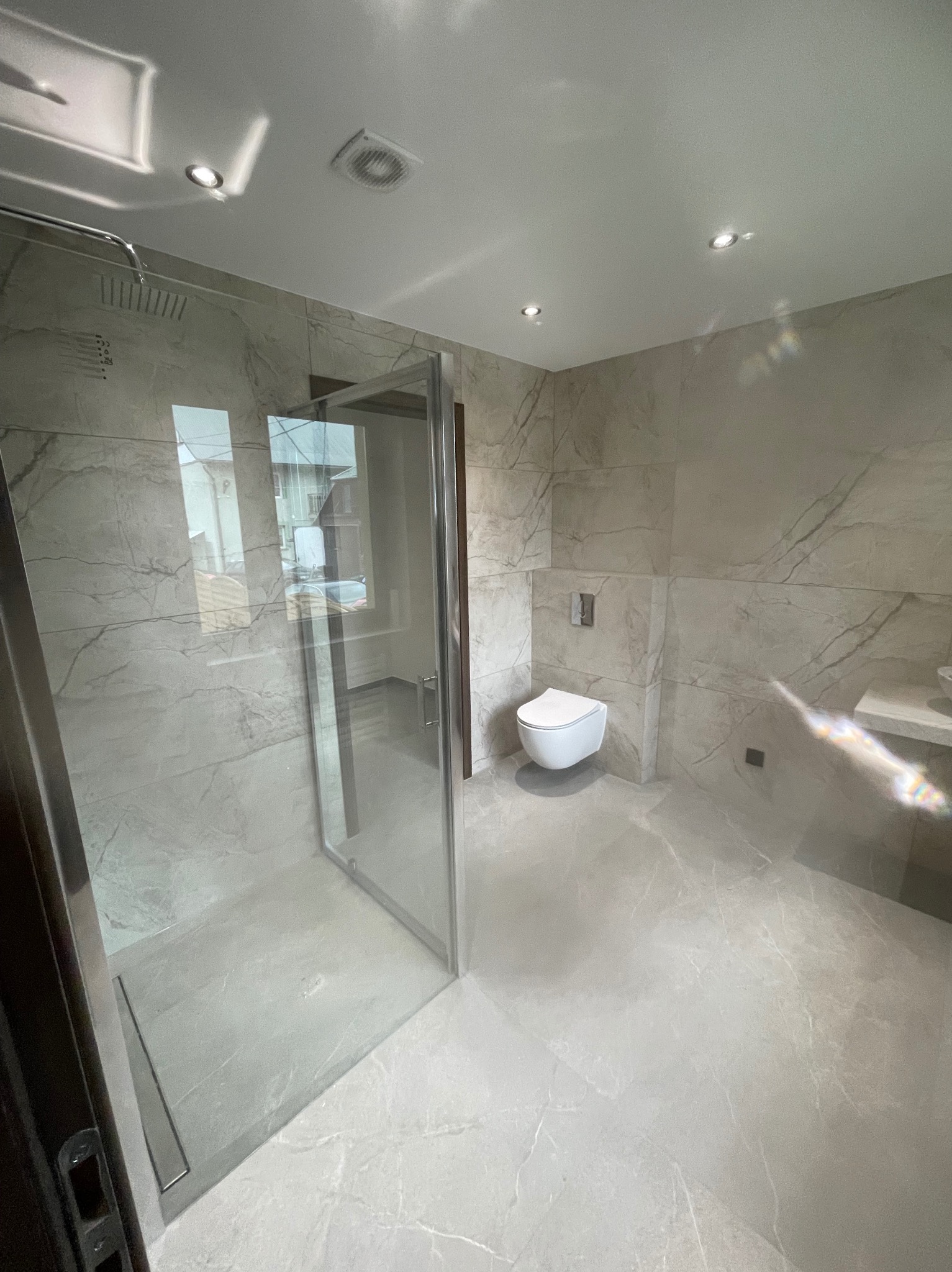 Nowoczesna łazienka z kabiną prysznicową typu walk-in, wykończona płytkami imitującymi marmur w odcieniach szarości. Podwieszana toaleta i minimalistyczna armatura.