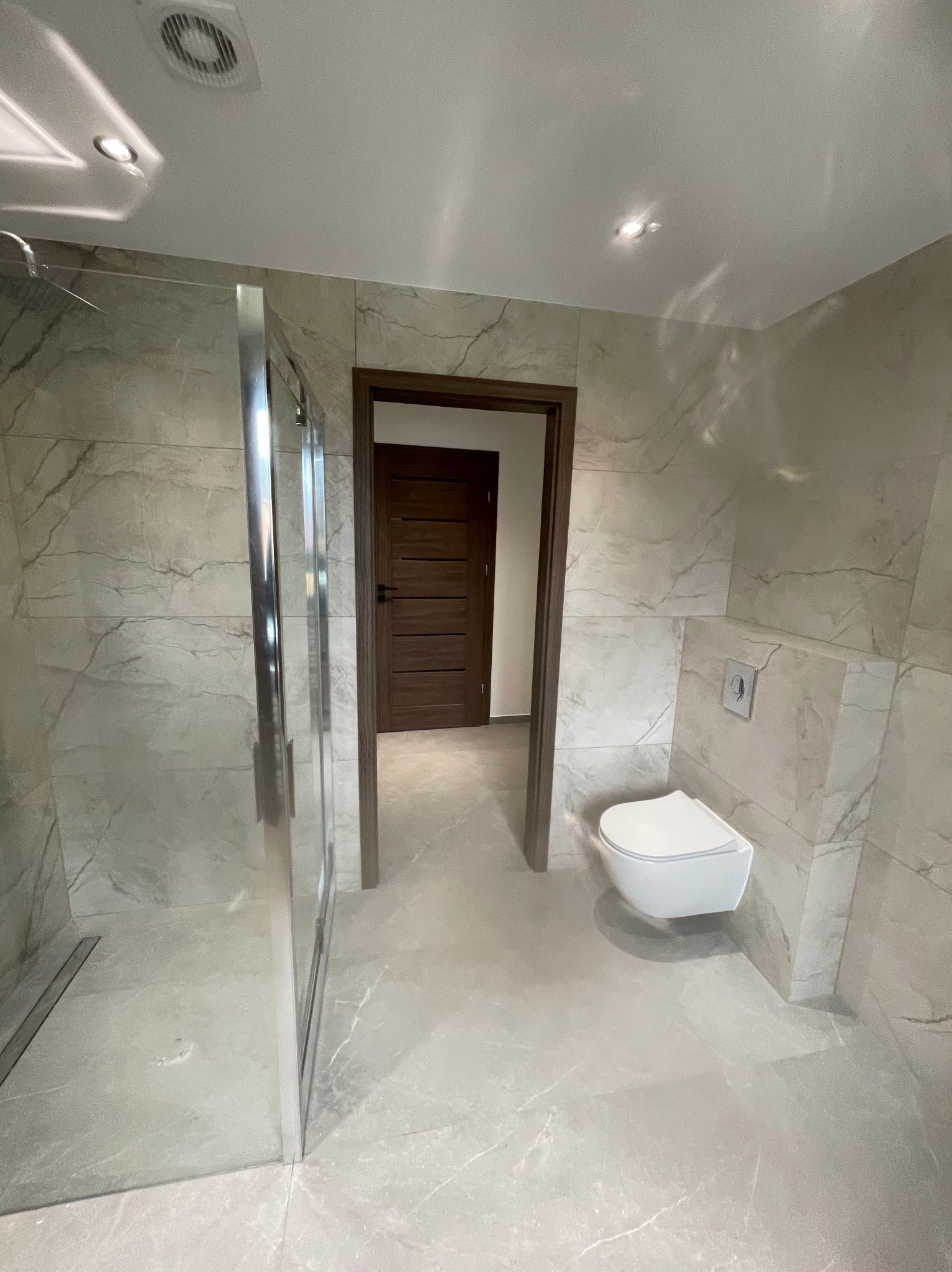 Wykończona łazienka w szarościach z kabiną prysznicową typu walk-in, podwieszaną toaletą i drzwiami z ciemnego drewna w tle. Płytki imitujące marmur na ścianach i podłodze.