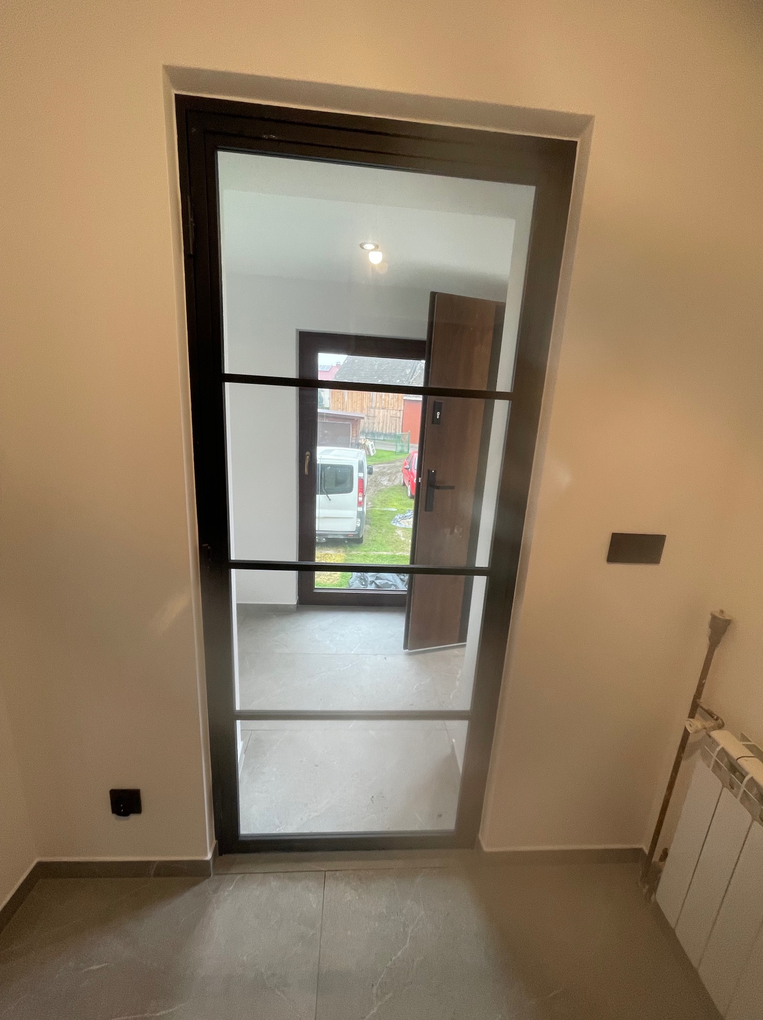 Nowoczesne drzwi wewnętrzne z czarną ramą i szprosami, widok na korytarz i otoczenie domu. Szare płytki podłogowe, świeżo wykończone ściany.