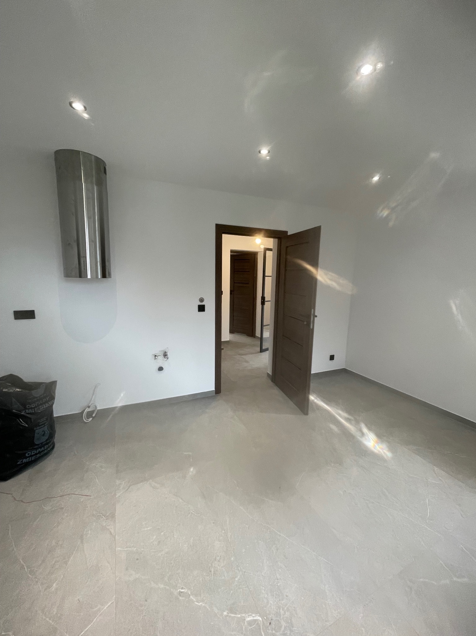 Jasne wnętrze po remoncie z szarą marmurową podłogą, otwartymi drzwiami i elementami instalacji na ścianie. Widoczny worek z odpadami budowlanymi.