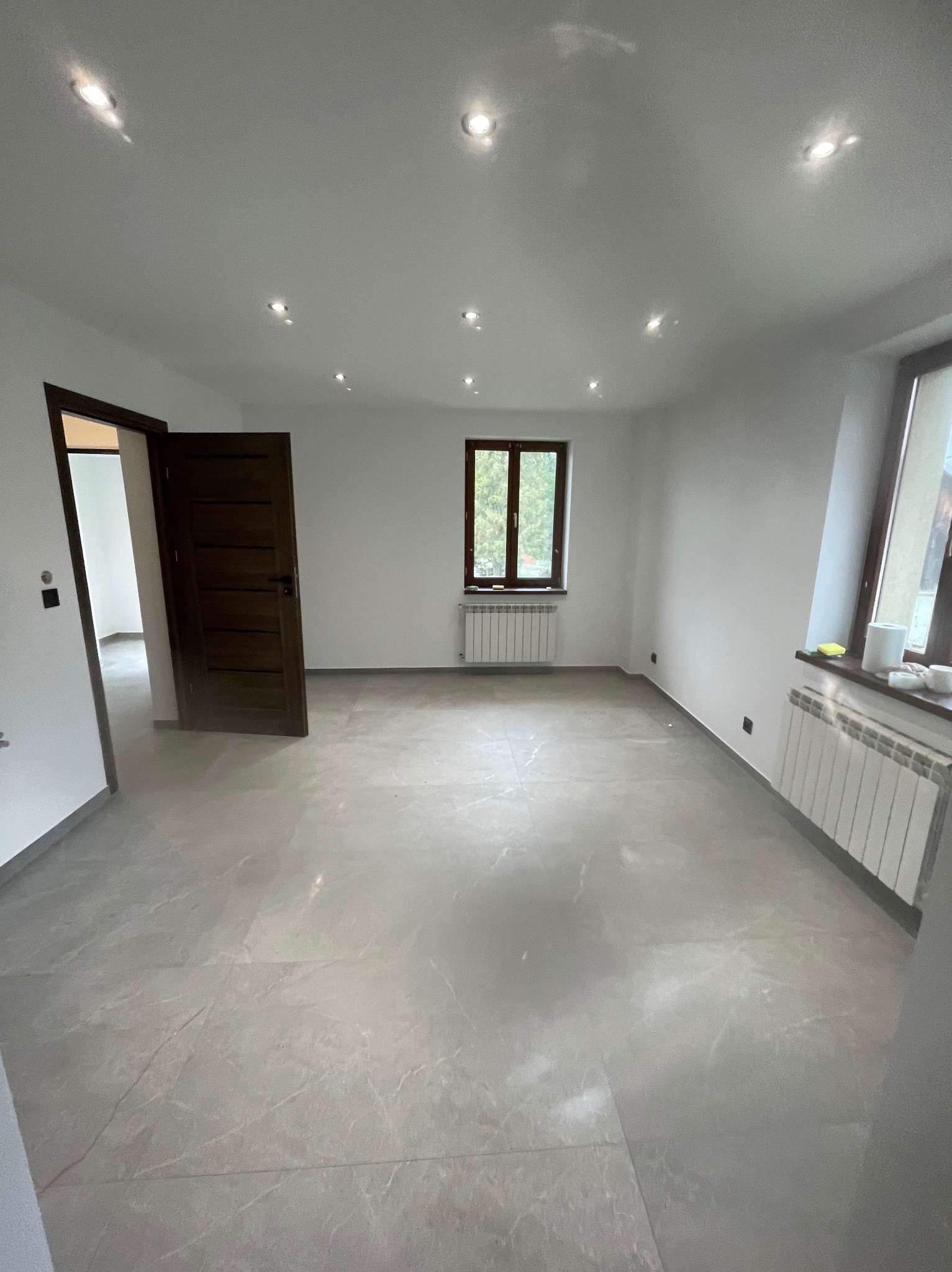 Jasne wnętrze po remoncie: szare płytki podłogowe, białe ściany, okna z ciemnego drewna i grzejniki. Widoczne drzwi wewnętrzne z ciemnego drewna i oświetlenie sufitowe.