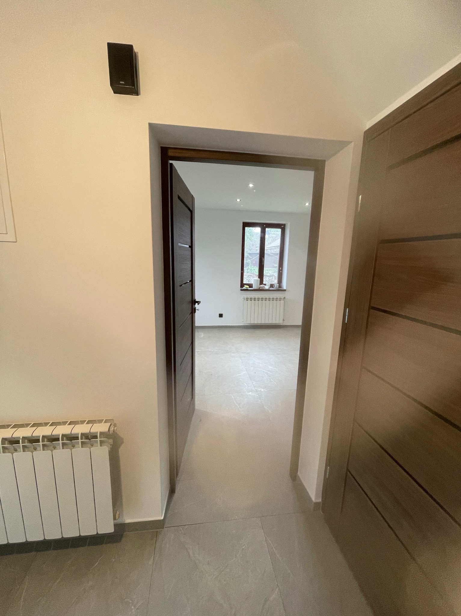 Wykończone wnętrze z widokiem na pokój przez otwarte, drewniane drzwi. Szare płytki podłogowe, białe ściany i grzejnik. Okno w oddali.