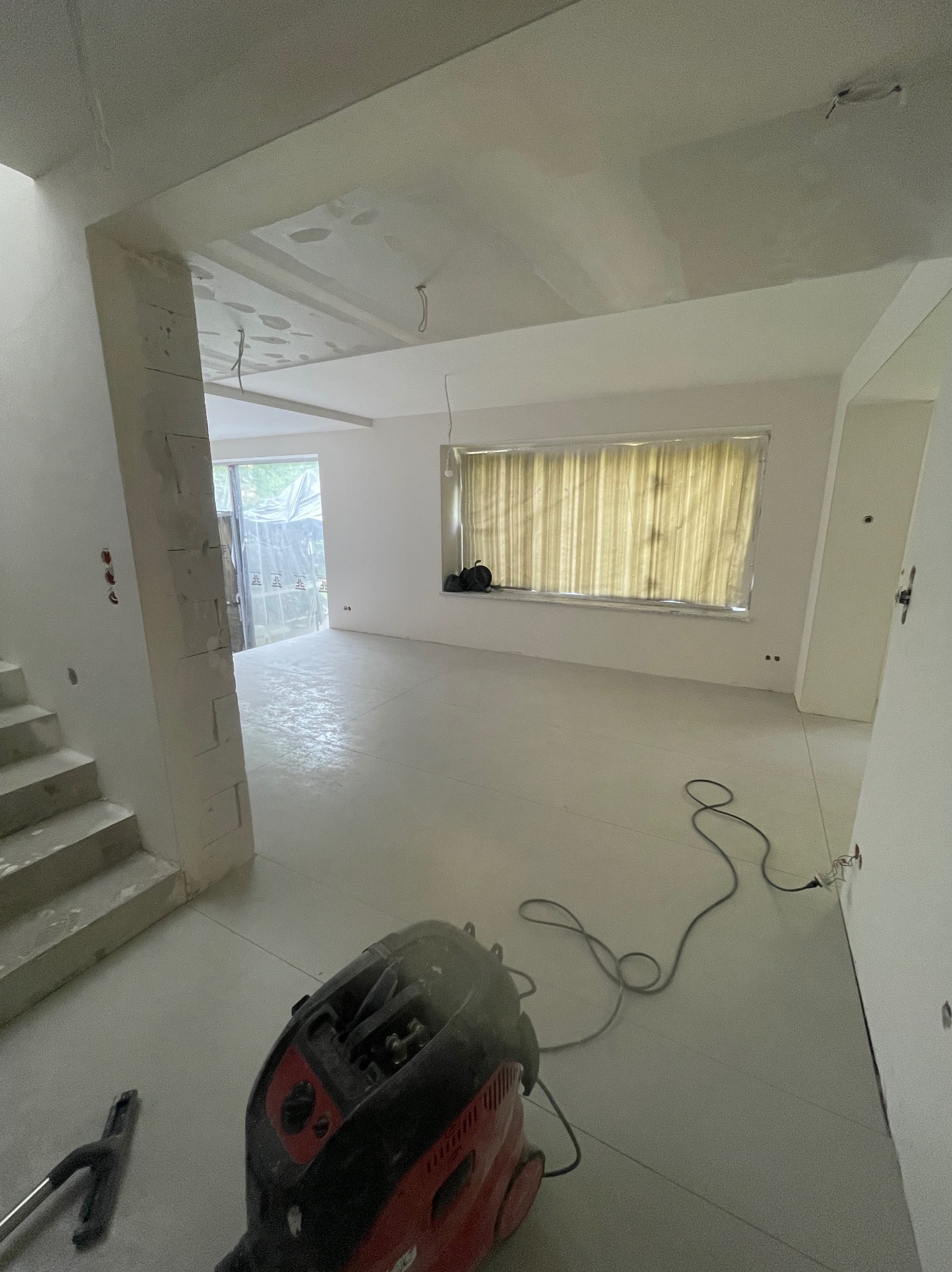 Wnętrze po szpachlowaniu i gładziowaniu, z widocznym odkurzaczem przemysłowym i schodami. Ściany i sufit przygotowane do malowania, okno zasłonięte folią.